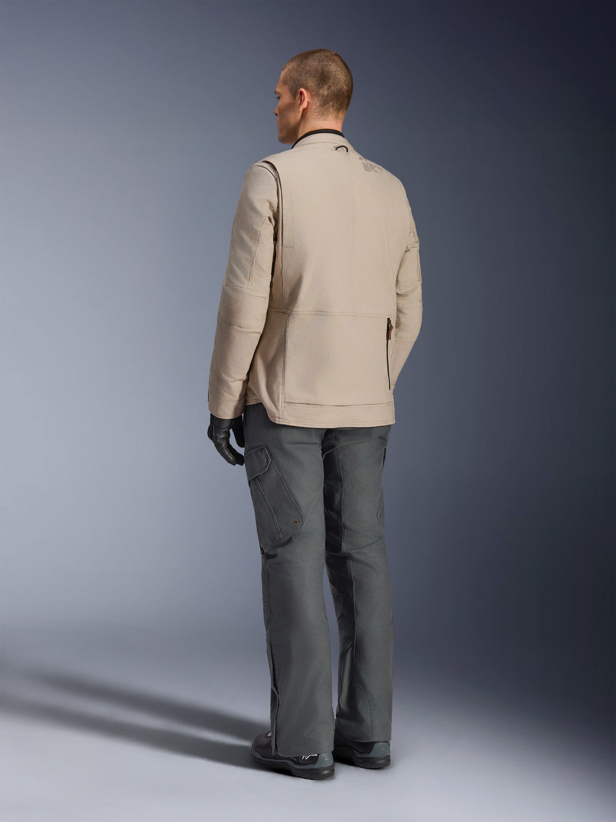 Alpinestars Borrego Drystar® Jacke in Warm Gray, Rückansicht mit robuster gewachster Canvas-Struktur, Erweiterungspanels an den hinteren Schultern und großer Reißverschlusstasche am Rücken, getragen von einem Model mit anthrazitgrauer Adventure-Hose und schwarzen Handschuhen.