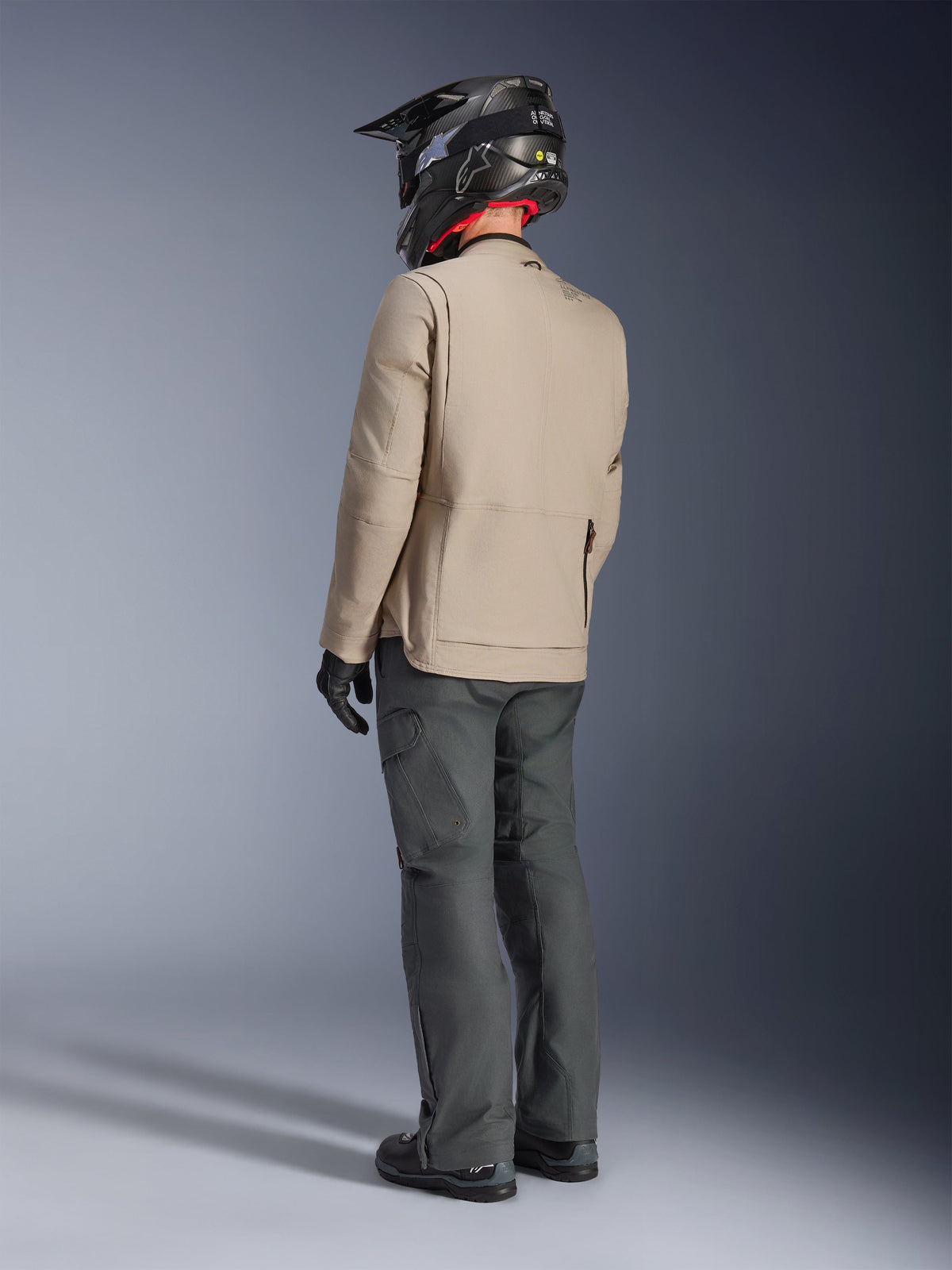 Alpinestars Borrego Drystar® Jacke, Abenteuer-Motorradjacke in Warm Gray, Rückansicht eines Fahrers mit dunkelgrauer Hose und schwarzem Helm, robuster gewachster Canvas-Look, funktionales Design