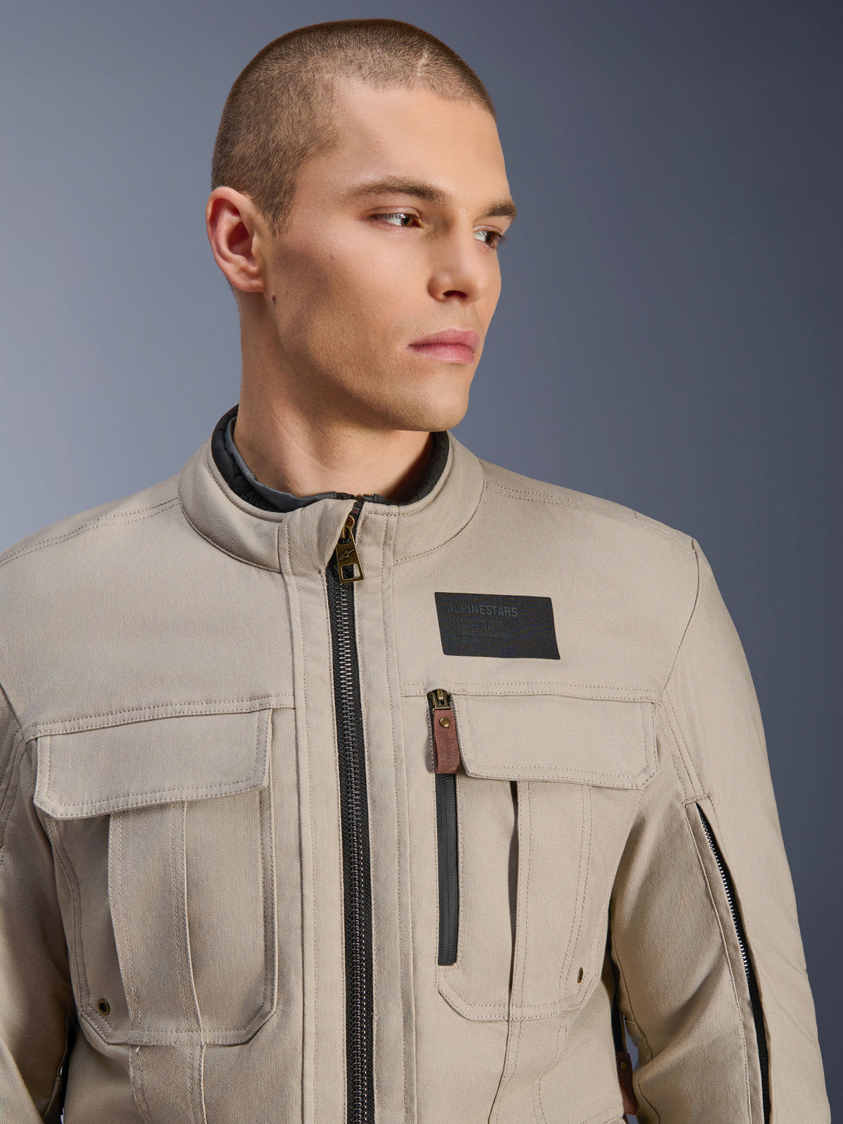 Männliches Model trägt die Alpinestars Borrego Drystar® Jacke in Warm Gray, Adventure-Motorradjacke mit klassischem gewachstem Canvas-Look, funktionalen Cargo-Brusttaschen und einem schwarzen Alpinestars Logo-Patch.