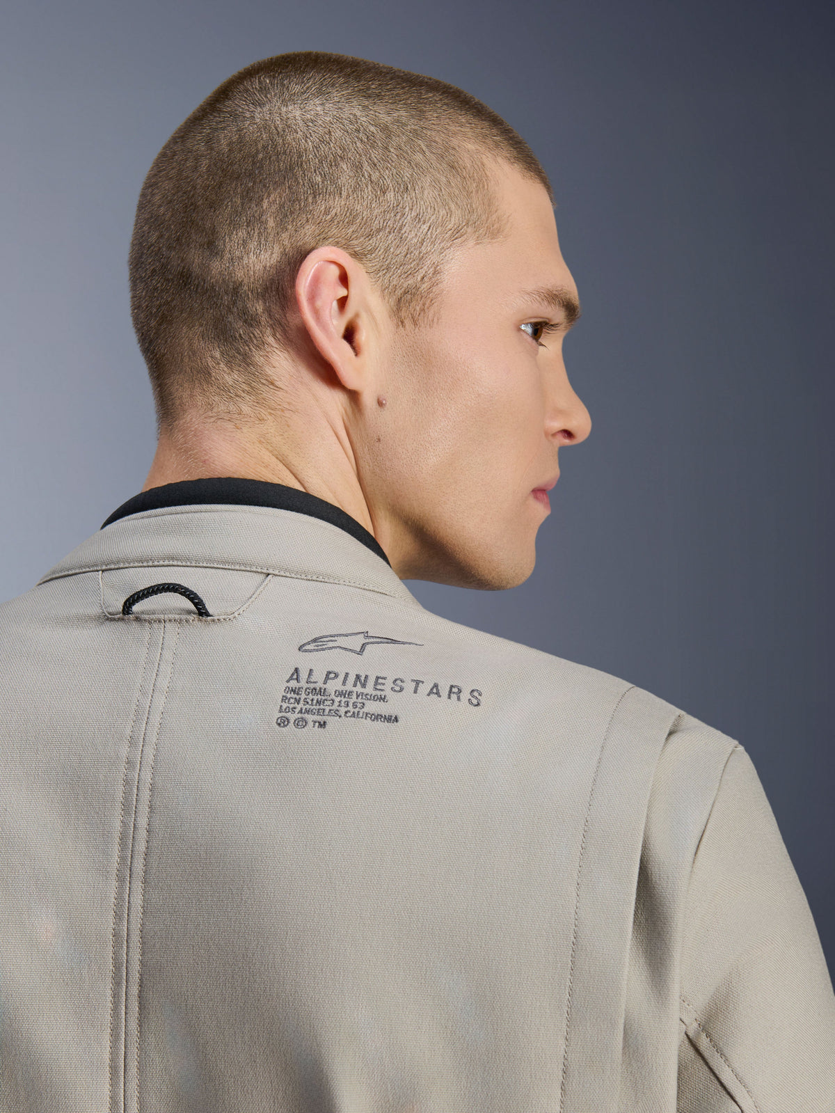 Alpinestars Borrego Drystar® Jacke in Warm Gray, Nahaufnahme der hinteren Schulter, texturiertes, gewachstes Canvas-Material, mit schwarzem Alpinestars-Logo und Heritage-Schriftzug, am Model, Adventure-Motorradjacke