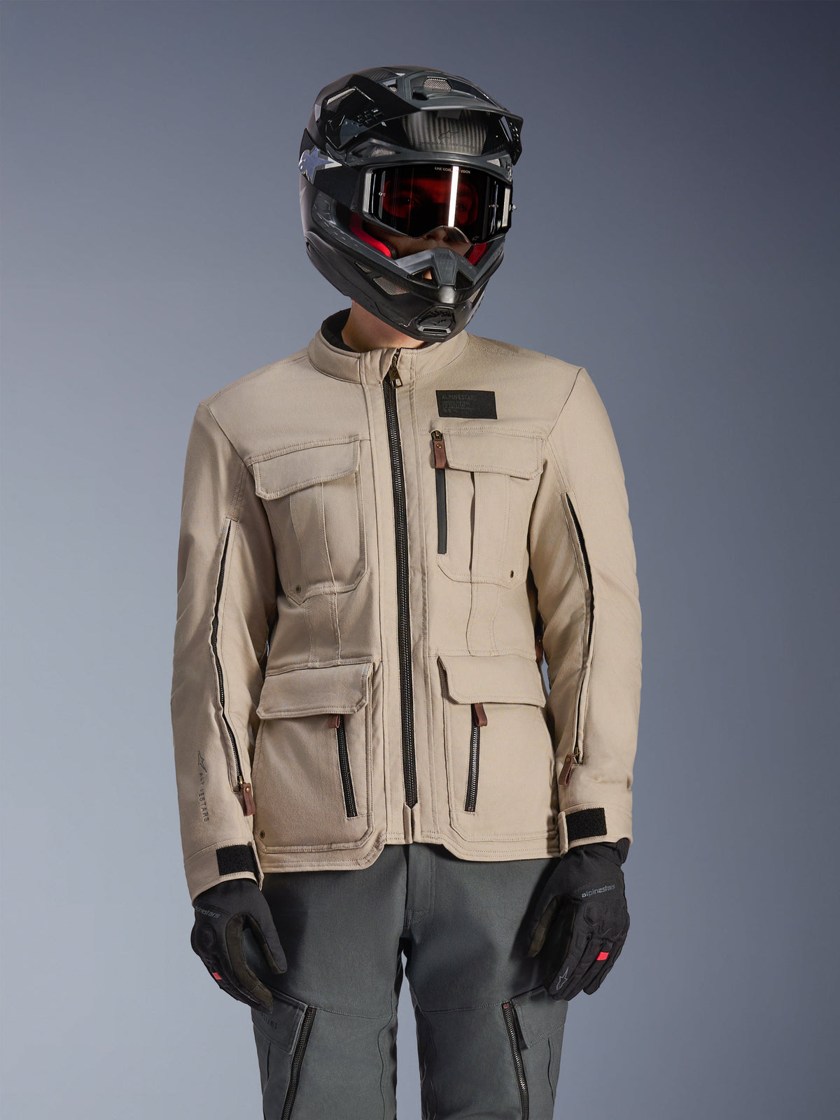 Alpinestars Borrego Drystar® Jacke, Adventure-Motorradjacke, Warmgrau, mit mehreren Cargo-Fronttaschen und schwarzen Reißverschlüssen, getragen mit schwarzem Helm, Schutzbrille und dunkelgrauer Motorradhose, robustes Textildesign für Touren