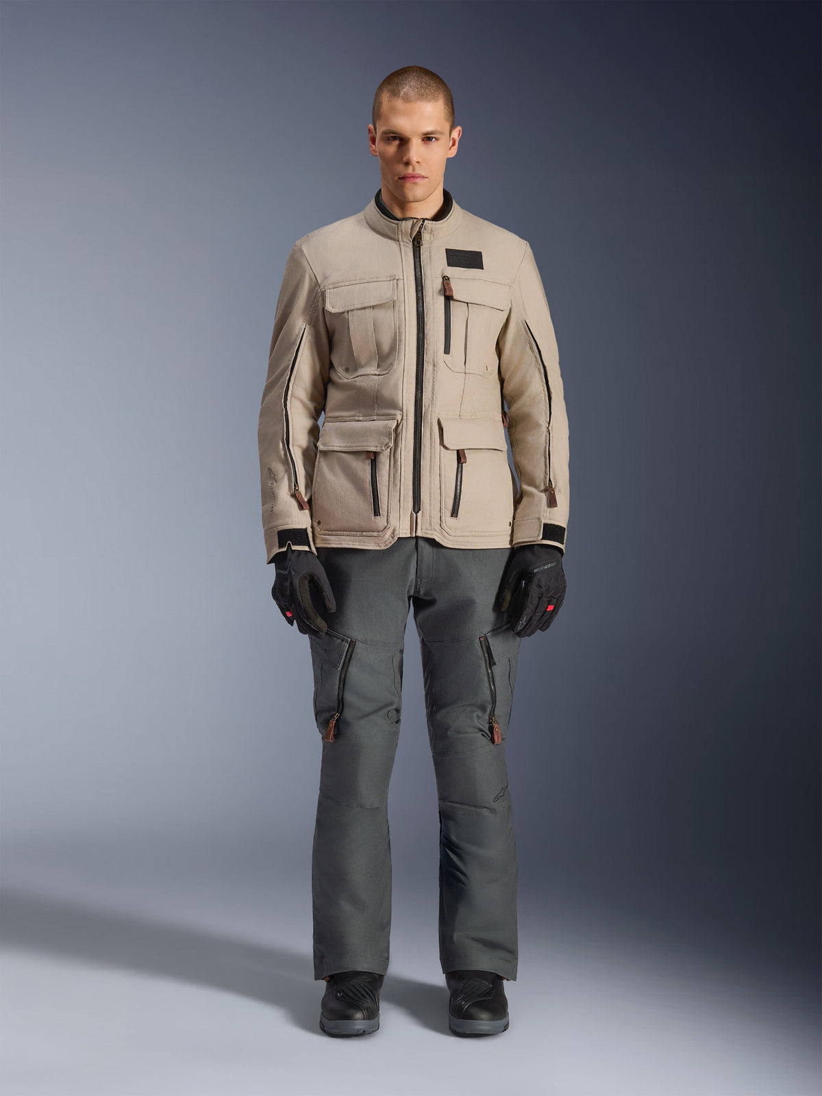 Alpinestars Borrego Drystar® Jacke, Adventure-Motorradjacke, Warm Gray, getragen von einem Modell mit dunkelgrauer Fahrhose und schwarzen Handschuhen, mit mehreren Cargo-Taschen und einer strapazierfähigen Textilkonstruktion im klassischen gewachsten Canvas-Look.