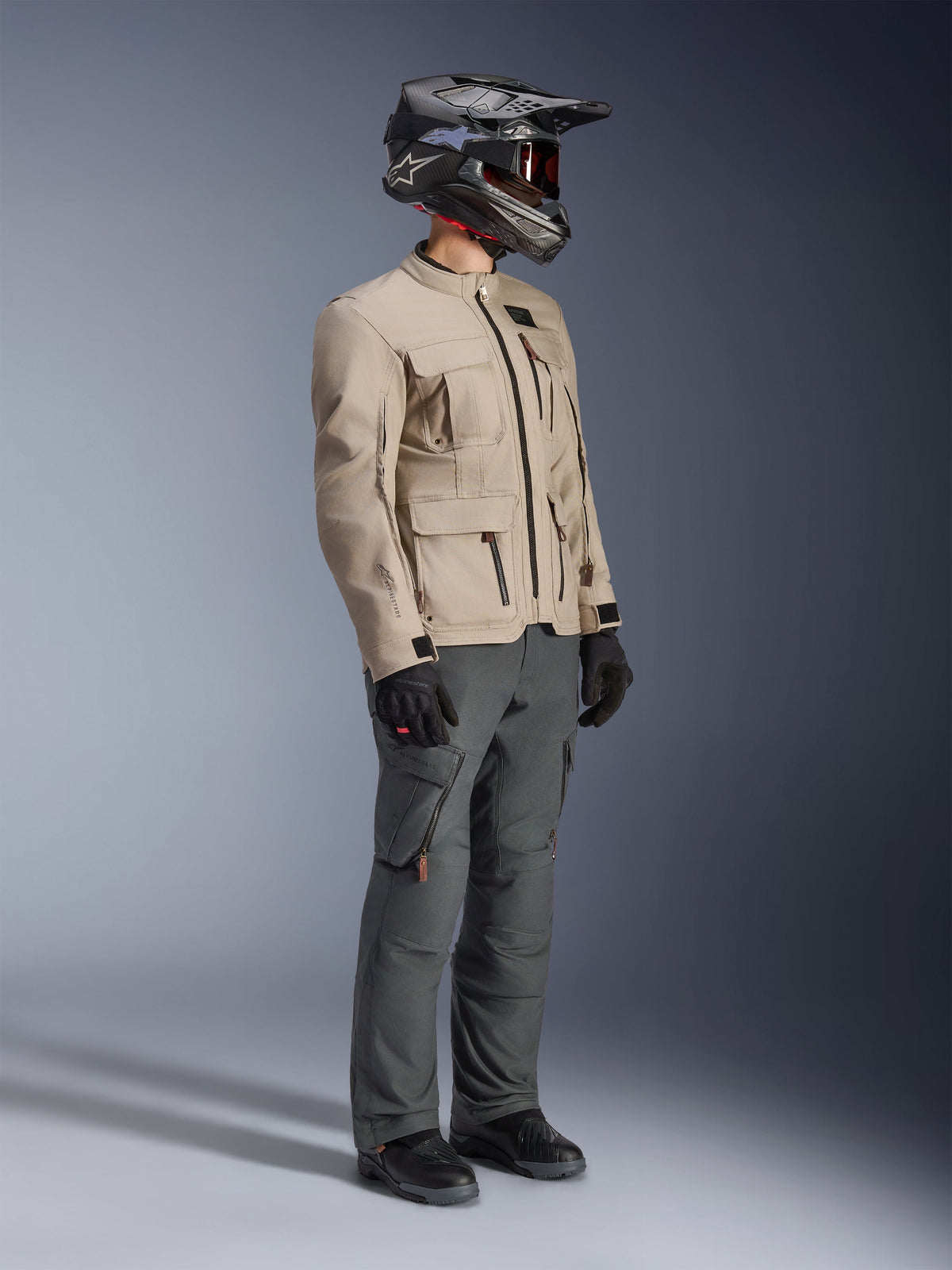 Motorradfahrer mit Alpinestars Borrego Drystar® Jacke in Warm Gray, Adventure-Motorradjacke mit gewachster Canvas-Struktur und Utility-Taschen, kombiniert mit dunkelgrauer Motorradhose, einem Carbon-Helm, schwarzen Handschuhen und schwarzen Stiefeln, Ganzkörper-Studioansicht eines kompletten Adventure-Touring-Outfits.