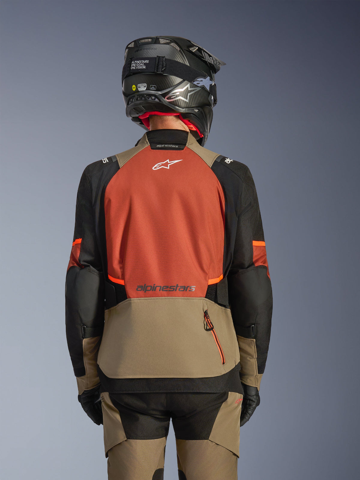 Rückansicht der Alpinestars Andes V4 Drystar® Jacke in Walnut Rust Black, Adventure-Tourenjacke mit einem großen rostfarbenen Rückenpanel und weißem Logo, walnussfarbenem unterem Bereich mit vertikaler Reißverschlusstasche, schwarzen Ärmeln mit orangefarbenen Akzenten, kombiniert mit passender Hose und einem schwarzen Helm für Fahrten bei jedem Wetter.