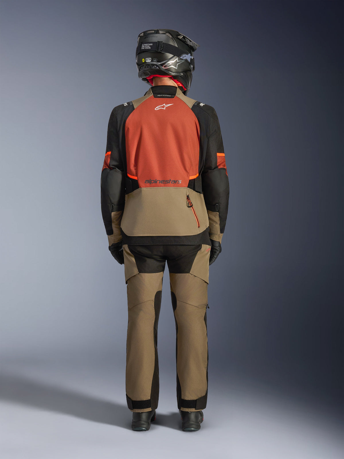 Rear view of a rider wearing the Alpinestars Andes V4 Drystar® Jacket in Walnut Rust Black, paired with matching walnut adventure pants and a black helmet, showcasing the jacket's rust-colored back panel, large rear storage pocket, and reflective logos.
