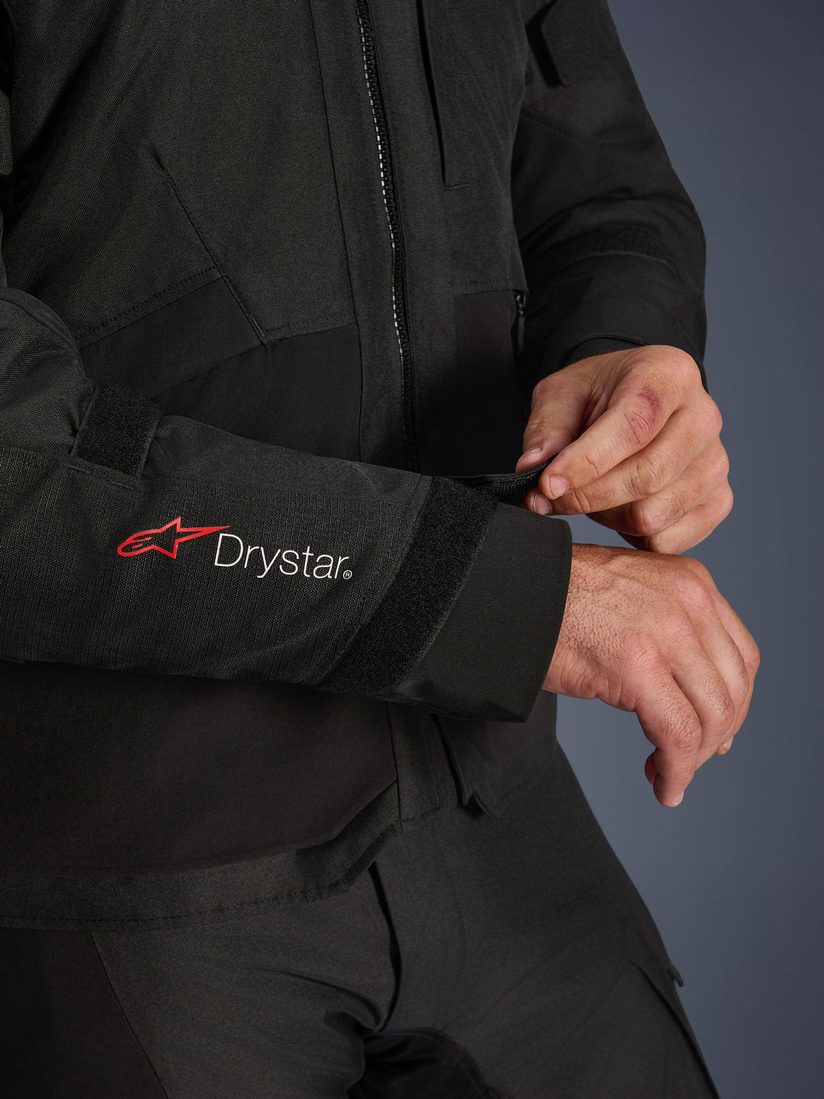 Alpinestars Andes V4 Drystar® Jacke, schwarze Textil-Motorradjacke, Nahaufnahme des Ärmels mit rotem Alpinestars-Logo und weißem Drystar®-Branding, Fahrer passt den Klettverschluss am Handgelenk für einen sicheren Sitz an, funktionales Detail für Adventure-Fahrten