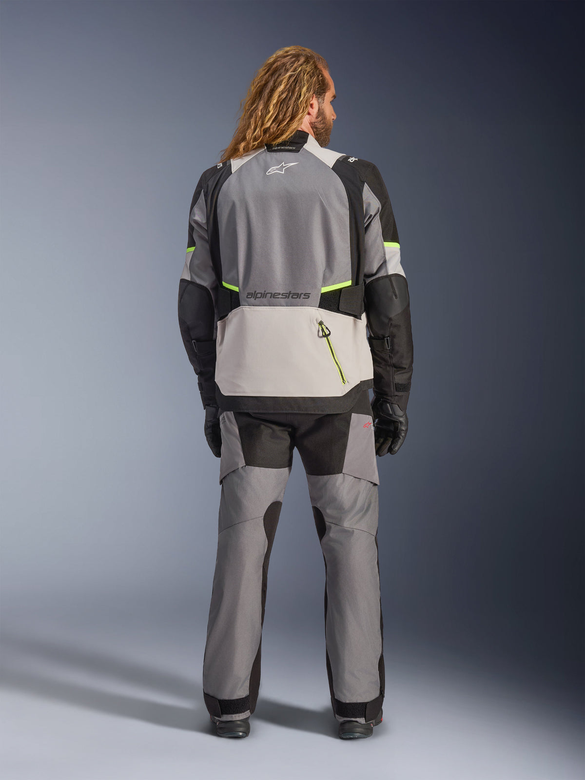 Alpinestars Andes V4 Drystar® Jacke, Adventure-Motorradjacke, Dunkelgrau und Eisgrau mit schwarzen und neongelben Akzenten, Rückansicht getragen mit passender Hose und schwarzen Handschuhen, große Rückentasche mit gelbem Reißverschluss, verstärkte Schulter- und Ellbogenpartien
