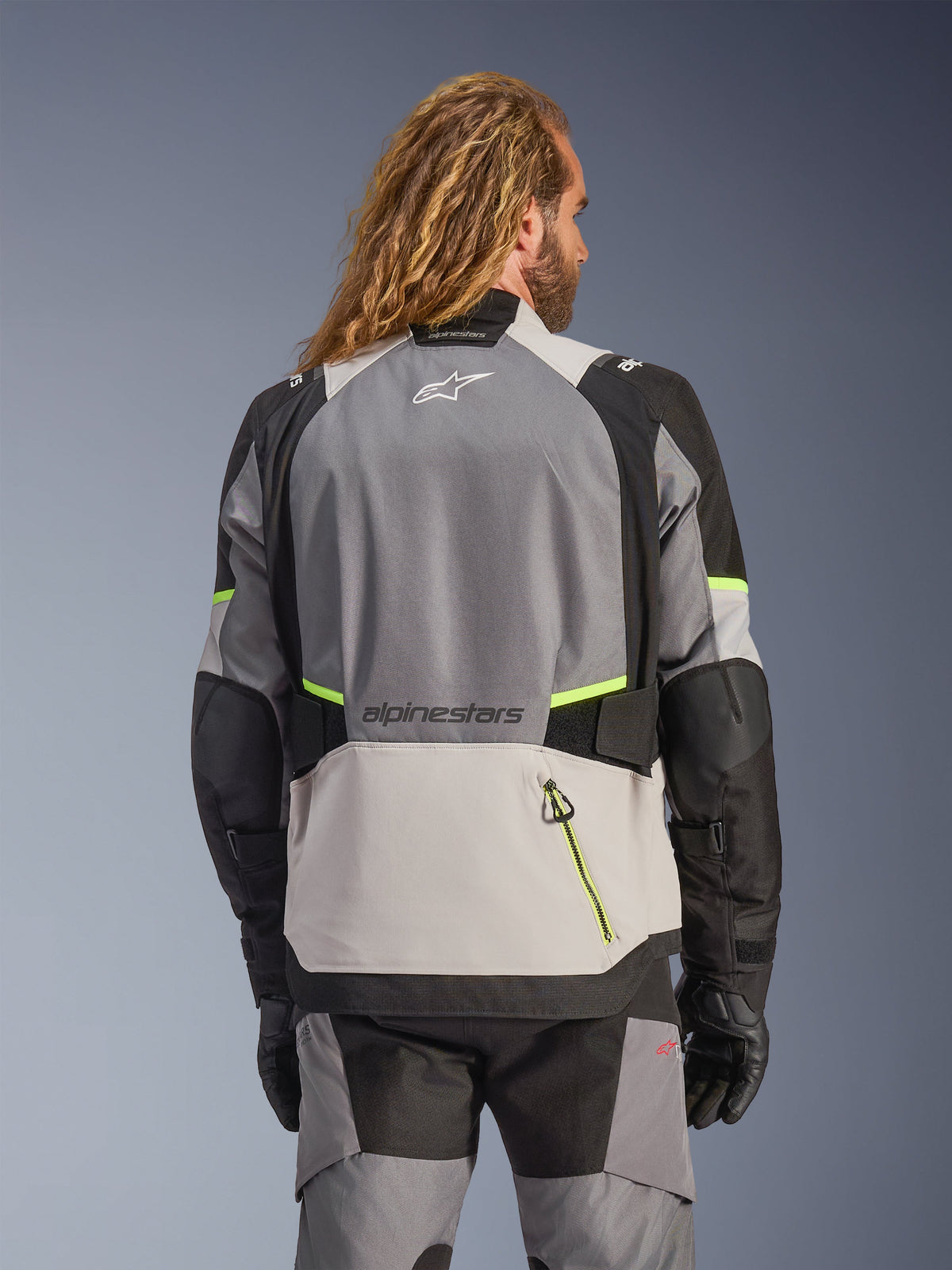 Alpinestars Andes V4 Drystar® Jacke, Rückansicht einer Adventure-Motorradjacke in Dunkelgrau, Eisgrau und Schwarz mit neongelben Akzenten, mit verstärkten Textileinsätzen und Alpinestars-Branding, getragen mit passender Motorradhose und schwarzen Handschuhen