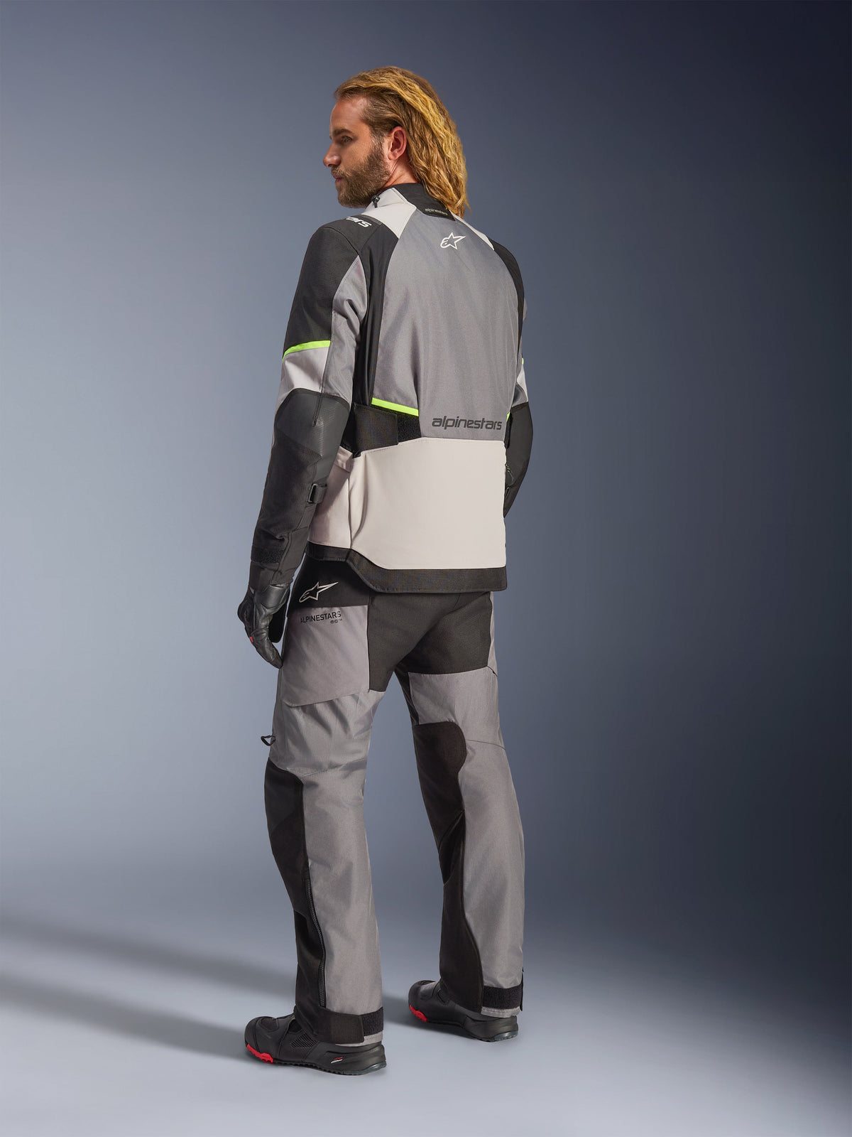 Alpinestars Andes V4 Drystar® Jacke, Dunkelgrau Ice-Grau Schwarz und Gelb Fluo, Rückansicht eines Fahrers mit passender Adventure-Tourenhose und Stiefeln, mit verstärkten grauen Textileinsätzen, neongelben Akzenten und Alpinestars-Branding, Allwetter-Motorradausrüstung.