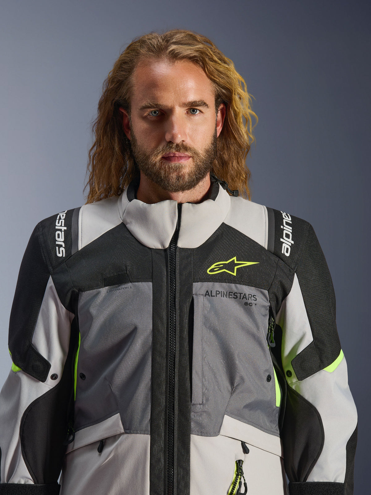 Alpinestars Andes V4 Drystar® Jacke, Dunkelgrau Ice Gray Schwarz und Gelb Fluo, getragen von einem Mann mit langem welligem Haar, technische Multi-Gewebe-Konstruktion, Alpinestars-Branding auf Brust und Schultern, hoher Kragen für Adventure-Fahrten