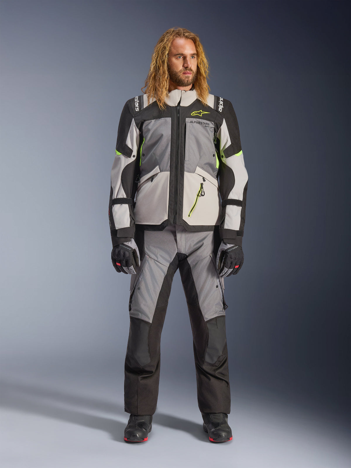 Männliches Model mit langen blonden Haaren in der Andes V4 Drystar® Jacke in Dunkelgrau, Eisgrau, Schwarz und Gelb Fluo, kombiniert mit einer passenden Adventure-Motorradhose und schwarzen Stiefeln, komplette technische Motorradausrüstung für Allwetter-Touren.
