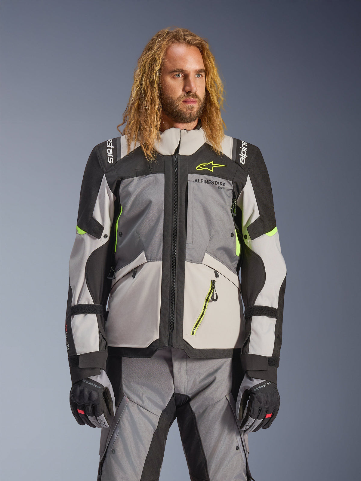 Alpinestars Andes V4 Drystar Jacke, Adventure-Touring-Motorradjacke, Dunkelgrau und Eisgrau mit neongelben Akzenten, getragen von einem Model mit passender Hose und Handschuhen, Multi-Pocket-Textildesign für Fahrten bei jedem Wetter