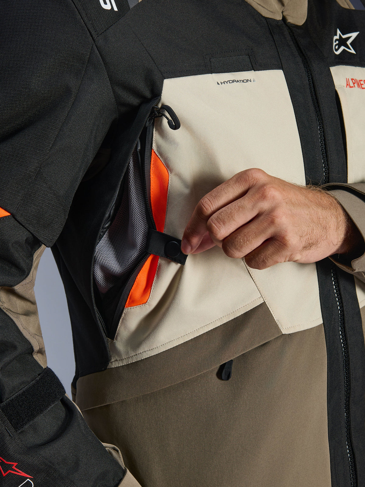 Alpinestars Andes V4 Drystar® Jacke Detailaufnahme, Textil in Walnut Mastic Black, Nahaufnahme einer Hand, die das Belüftungspanel an der Brust öffnet und ein leuchtend orangefarbenes Mesh-Gewebe freilegt, direktes Belüftungssystem für Adventure-Fahrten