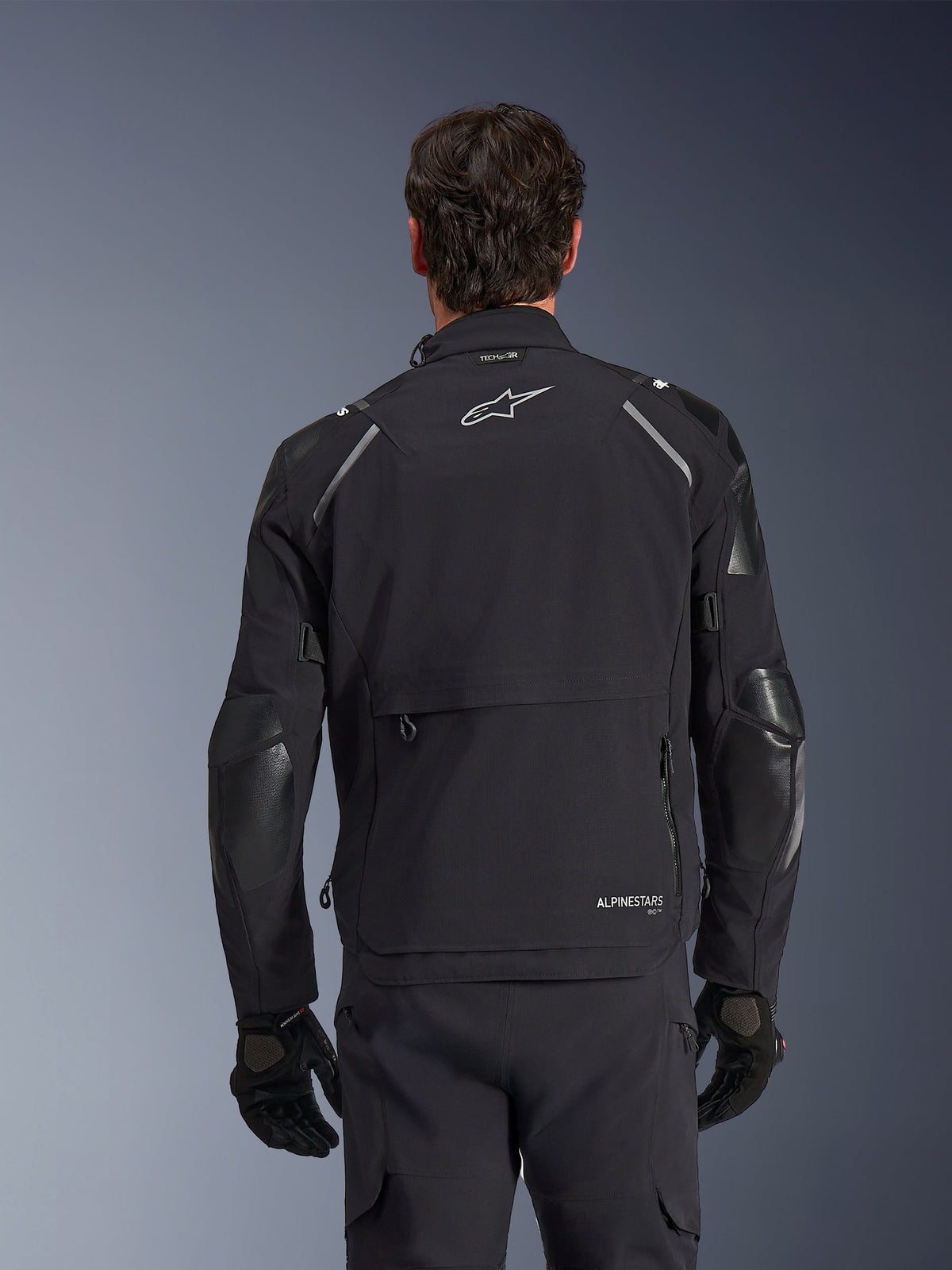 Alpinestars Andes Pro Drystar® XF Jacke, Schwarz und Dunkelgrau, Rückansicht an einem männlichen Modell, mit einem großen hinteren Staufach, reflektierenden Schulterakzenten und Logo-Branding, kombiniert mit schwarzen Handschuhen und einer Hose für Adventure-Touren.