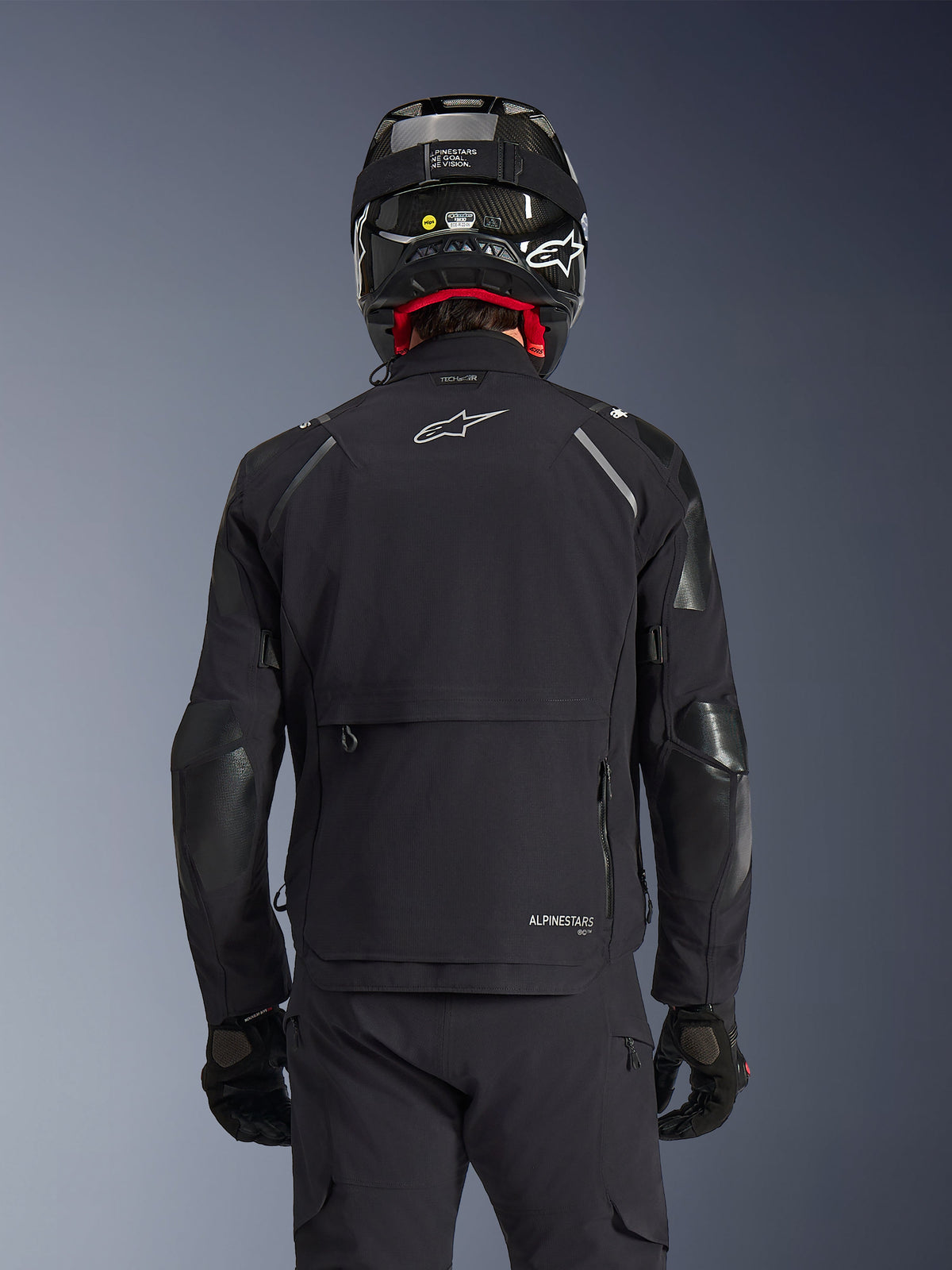 Alpinestars Andes Pro Drystar® XF Jacke in Schwarz, Rückansicht an einem Fahrer mit Carbonfaser-Helm, mit großem hinterem Staufach und weißem Alpinestars-Logo, technische Ganzjahres-Adventure-Motorradausrüstung.