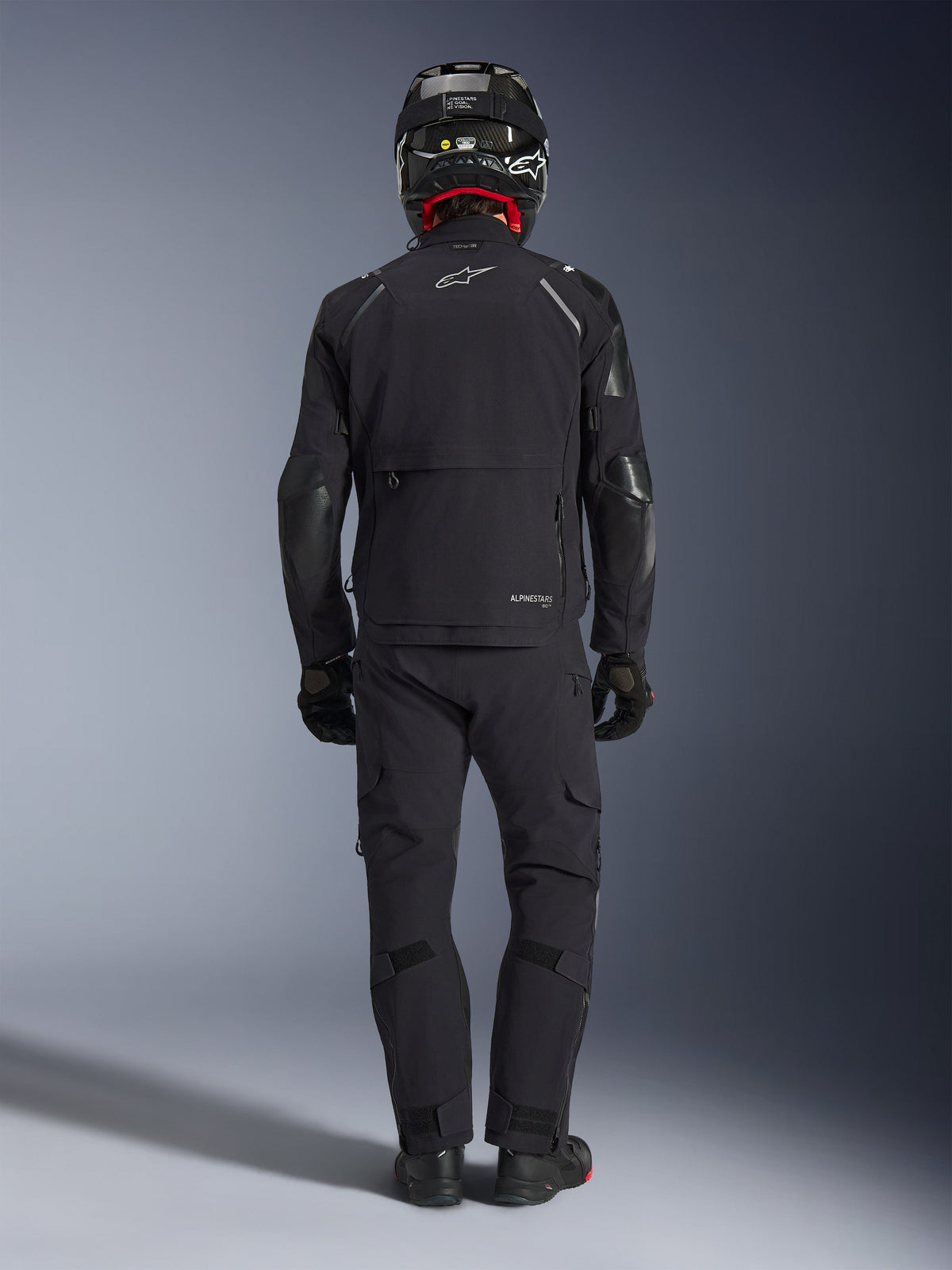 Alpinestars Andes Pro Drystar® XF Jacke in Schwarz und Dunkelgrau, Rückansicht eines Fahrers in vollständiger Adventure-Touring-Ausrüstung inklusive passender Hose und Carbonfaser-Helm, mit einer großen Cargo-Rückentasche und reflektierendem Alpinestars-Branding auf dem Rücken.