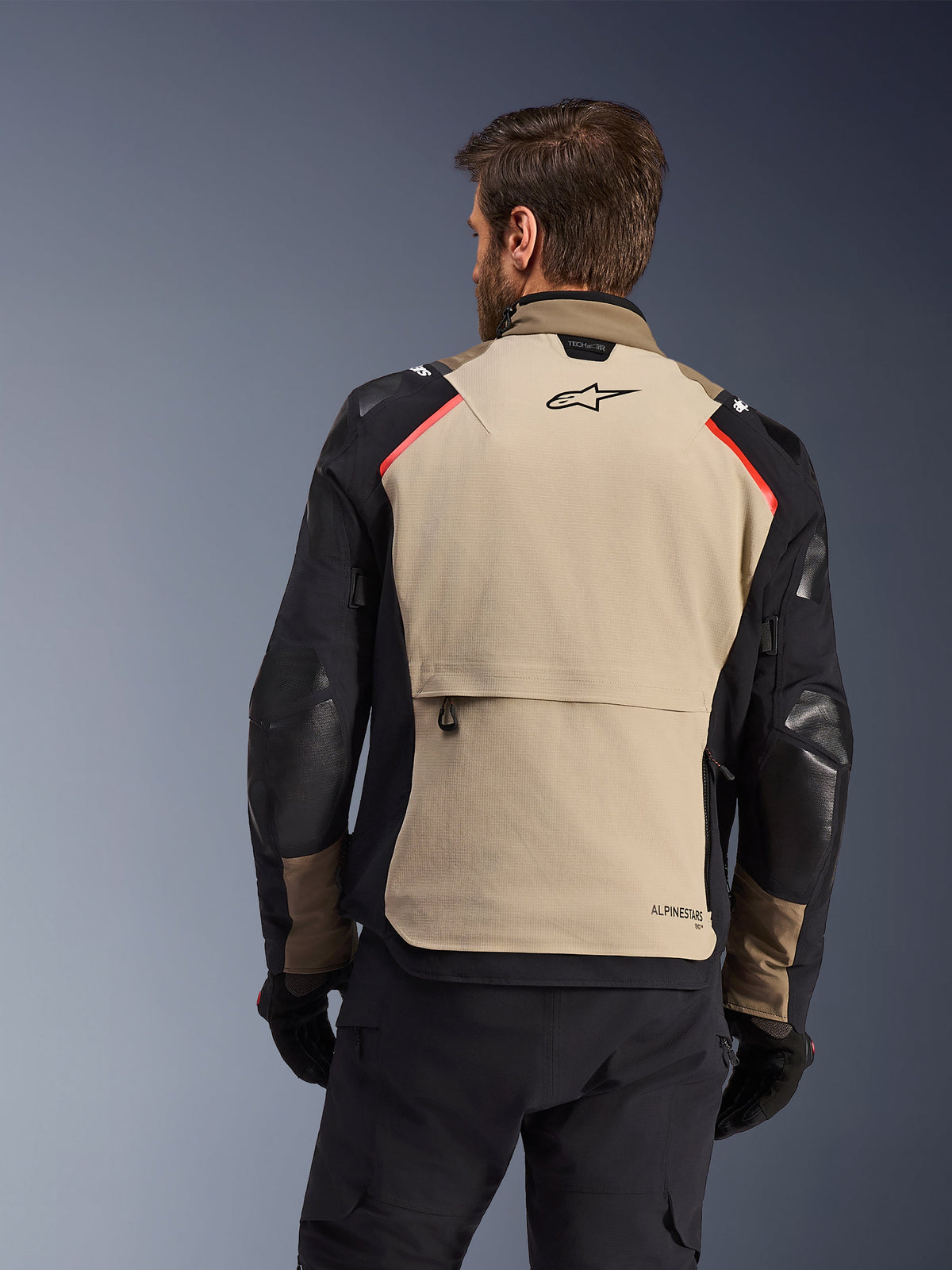 Alpinestars Andes Pro Drystar® XF Jacke, Rückansicht, Walnut und Aluminum, großes hellbraunes Rückenpanel mit schwarzem Alpinestars-Logo und roten Schulterakzenten, große Cargo-Rückentasche, getragen mit schwarzer Hose und Handschuhen, Adventure-Touren-Motorradausrüstung
