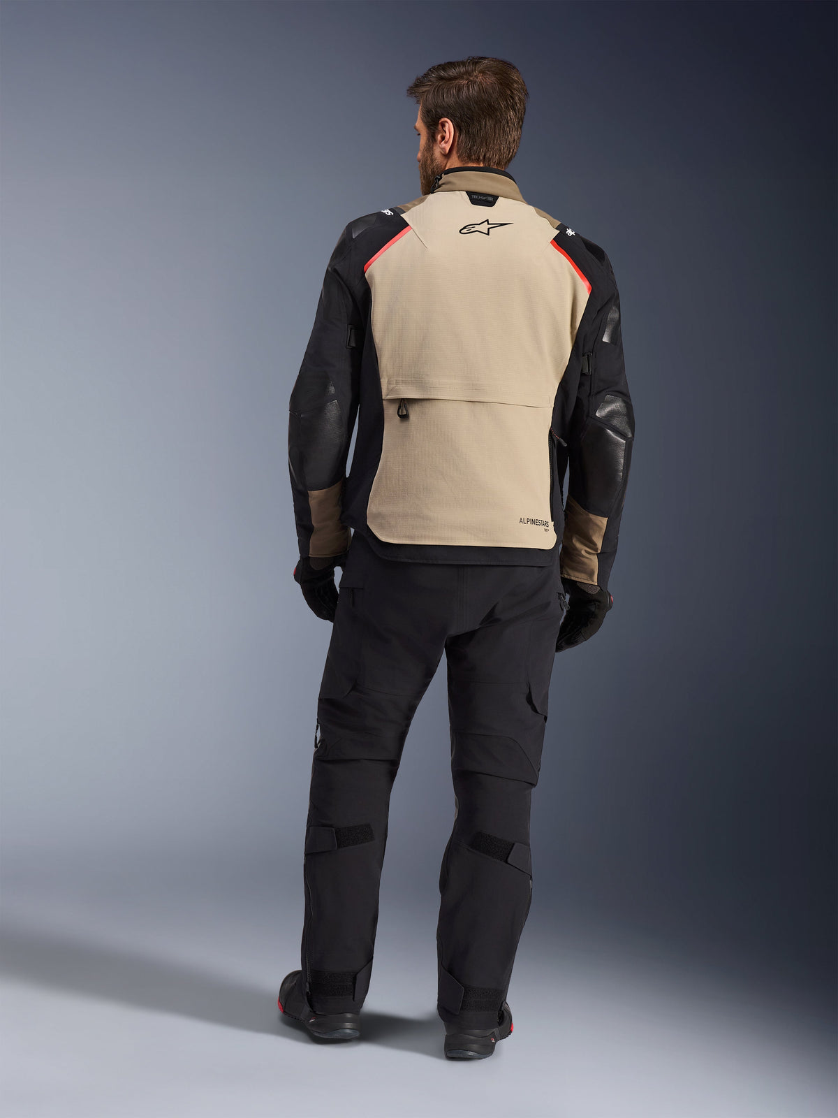 Alpinestars Andes Pro Drystar® XF Jacke, Walnut Aluminum, Rückansicht, Motorrad-Adventure-Jacke mit schwarzen Ärmeln und roten Schulterakzenten, getragen mit schwarzer Motorradhose und Handschuhen.
