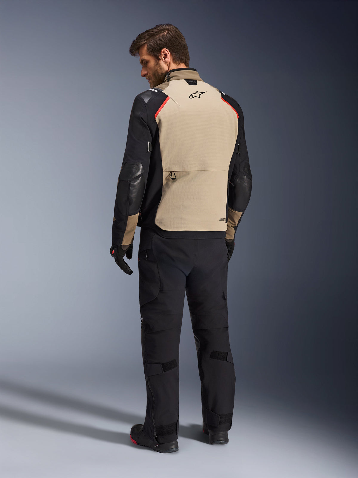 Alpinestars Andes Pro Drystar® XF Jacke, Motorradjacke, Walnuss und Schwarz mit roten Akzenten, Rückansicht am Modell, Adventure-Touring-Design mit großer Stautasche auf der Rückseite, getragen mit schwarzer Motorradhose und Handschuhen.