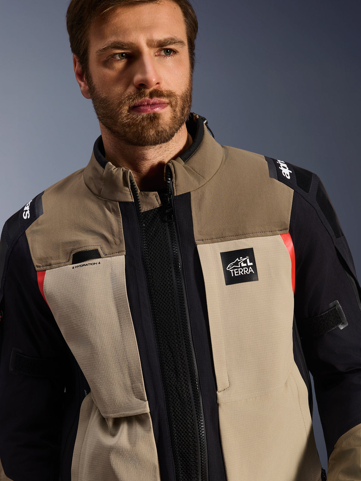 Alpinestars Andes Pro Drystar® XF Jacke in Walnut Aluminum, getragen von einem männlichen Model, Adventure-Motorradjacke mit einem mehrlagigen Textildesign in Braun, Hellgrau und Schwarz mit roten Akzenten, Alpinestars „TERRA“-Logo auf der Brust und sichtbaren technischen Details.