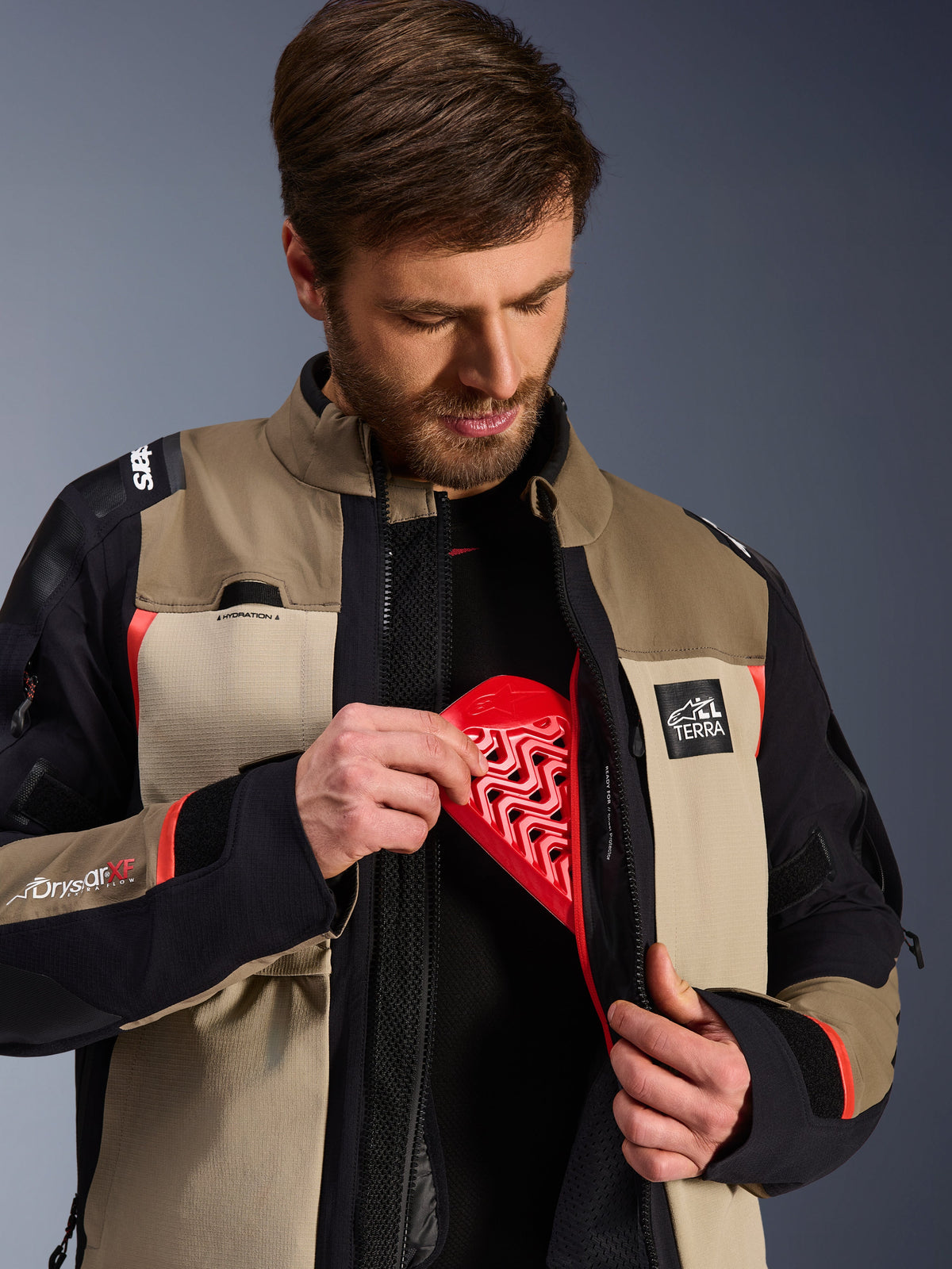 Alpinestars Andes Pro Drystar® XF Jacke in Walnut Aluminum, Modell demonstriert das Einsetzen des roten Nucleon Brustprotektors in die Innentasche, Adventure-Motorradjacke mit ALL TERRA Branding und Drystar XF Logos.