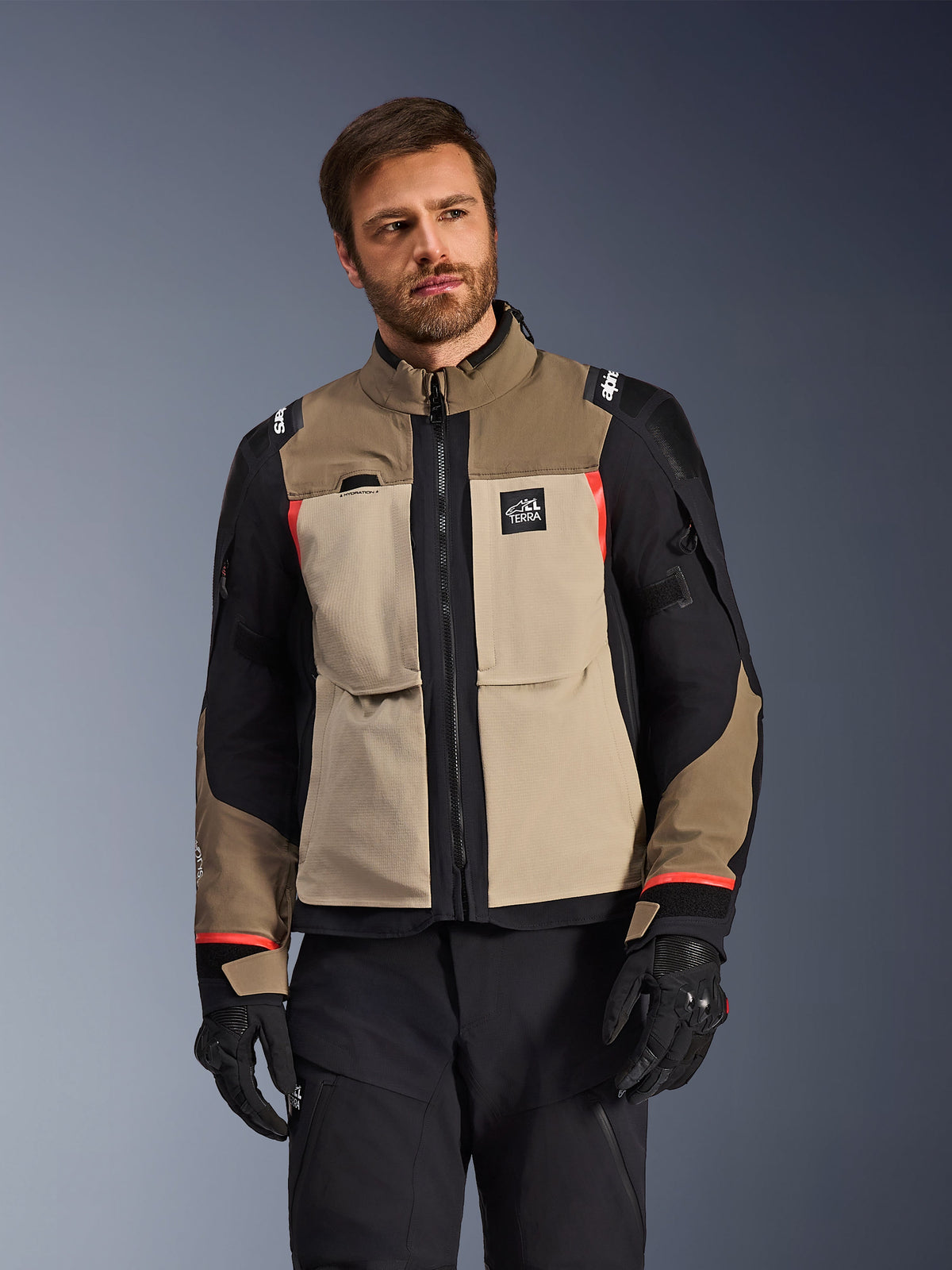 Alpinestars Andes Pro Drystar® XF Jacke, Walnut Aluminum, Motorrad-Adventure-Jacke mit hellbraunen und schwarzen Einsätzen und roten Akzenten, getragen mit schwarzer Motorradhose und Handschuhen, technische wasserdichte Tourenbekleidung