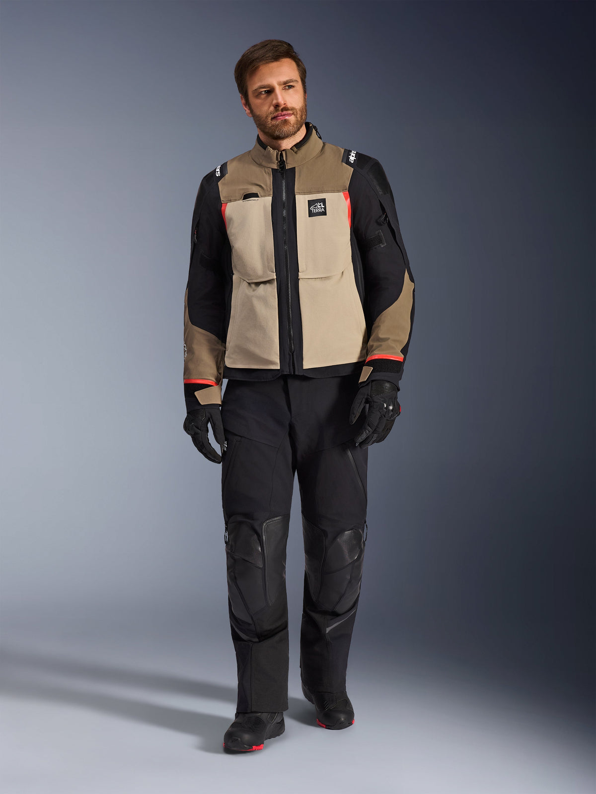 Alpinestars Andes Pro Drystar® XF Jacke in Walnut Aluminum, beige-schwarze Adventure-Touring-Jacke mit roten Akzenten, kombiniert mit schwarzer Motorradhose, Handschuhen und Stiefeln, Ganzkörper-Studioaufnahme für schützende Fahrerausrüstung.