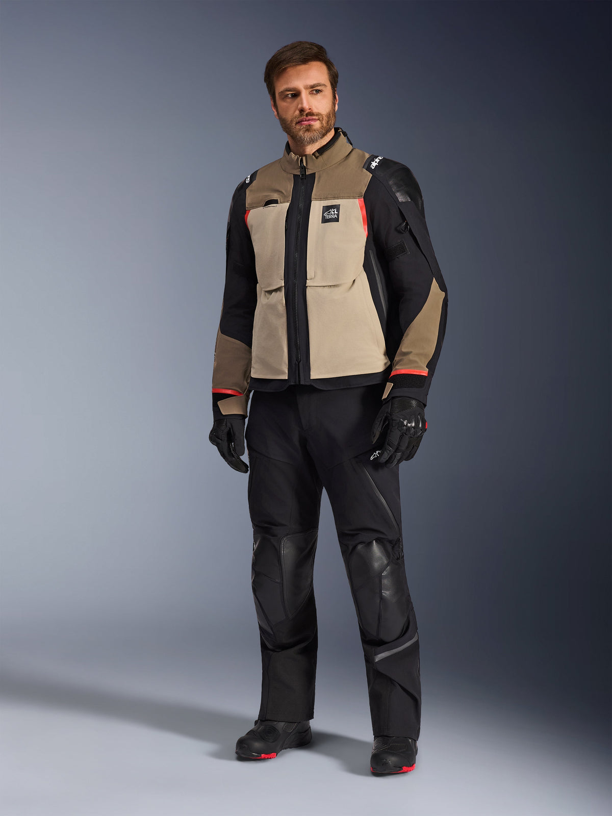 Ein männliches Model trägt die Alpinestars Andes Pro Drystar® XF Jacke in Walnut Aluminum, kombiniert mit einer schwarzen Motorradhose, Handschuhen und Stiefeln, mit einem mehrteiligen Design in Hellbraun und Schwarz mit roten Akzenten für Adventure-Touren.