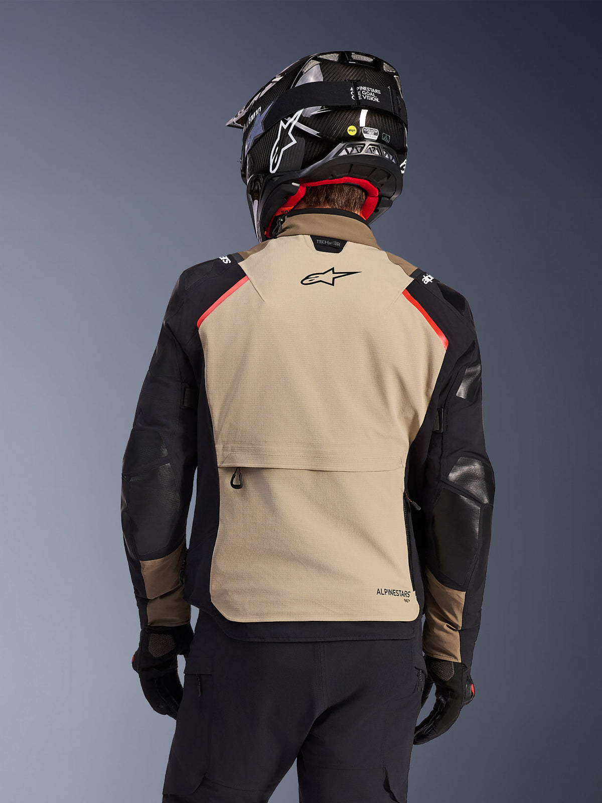 Alpinestars Andes Pro Drystar® XF Jacke in Walnut Aluminum, Rückansicht, tanfarbenes Rückenteil mit großer Cargo-Tasche, schwarze Ärmel mit verstärkten Einsätzen, Alpinestars-Logo auf dem oberen Rücken, kombiniert mit schwarzem Helm und Handschuhen für Adventure-Touren.