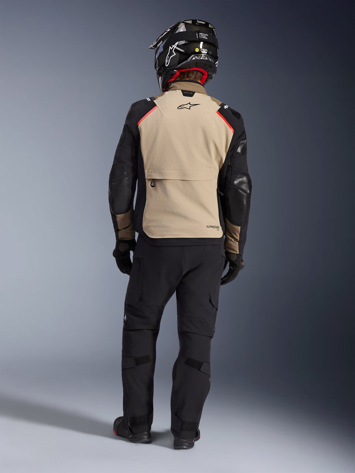 Rückansicht eines Fahrers mit der Alpinestars Andes Pro Drystar® XF Jacke in Walnut Aluminum, beigefarbenes Rückenteil mit schwarzen Ärmeln und roten Akzenten, kombiniert mit einer schwarzen technischen Adventure-Hose und einem Motorradhelm, hochwertige Allwetter-Tourenbekleidung.