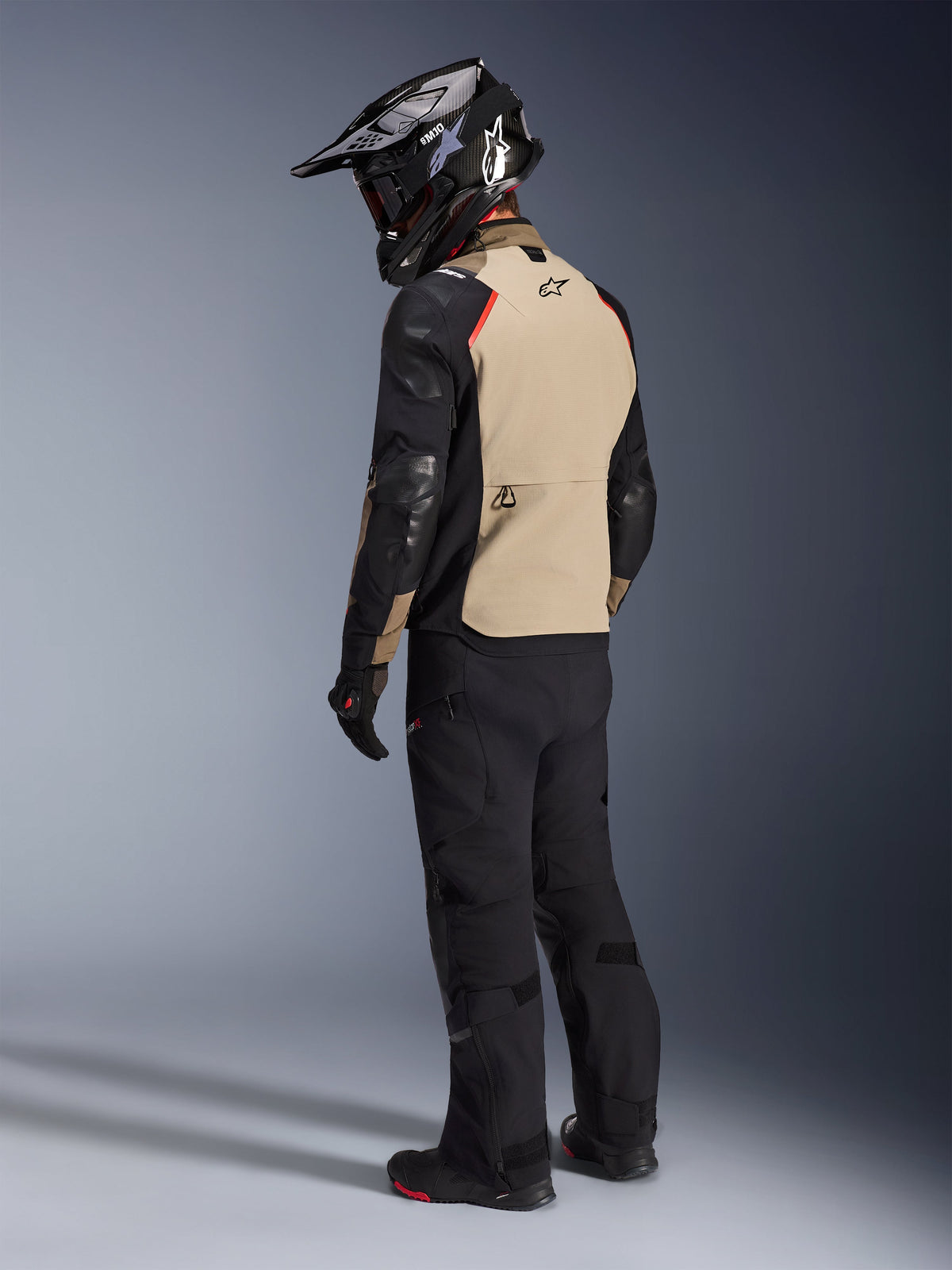 Motorradfahrer von hinten in der Alpinestars Andes Pro Drystar® XF Jacke in Walnut Aluminum, beige Rückenpartie mit schwarzen Ärmeln und roten Akzenten, schwarzer Adventure-Helm, schwarze Motorradhose und schwarze Stiefel, professionelle Adventure-Tourenausrüstung.