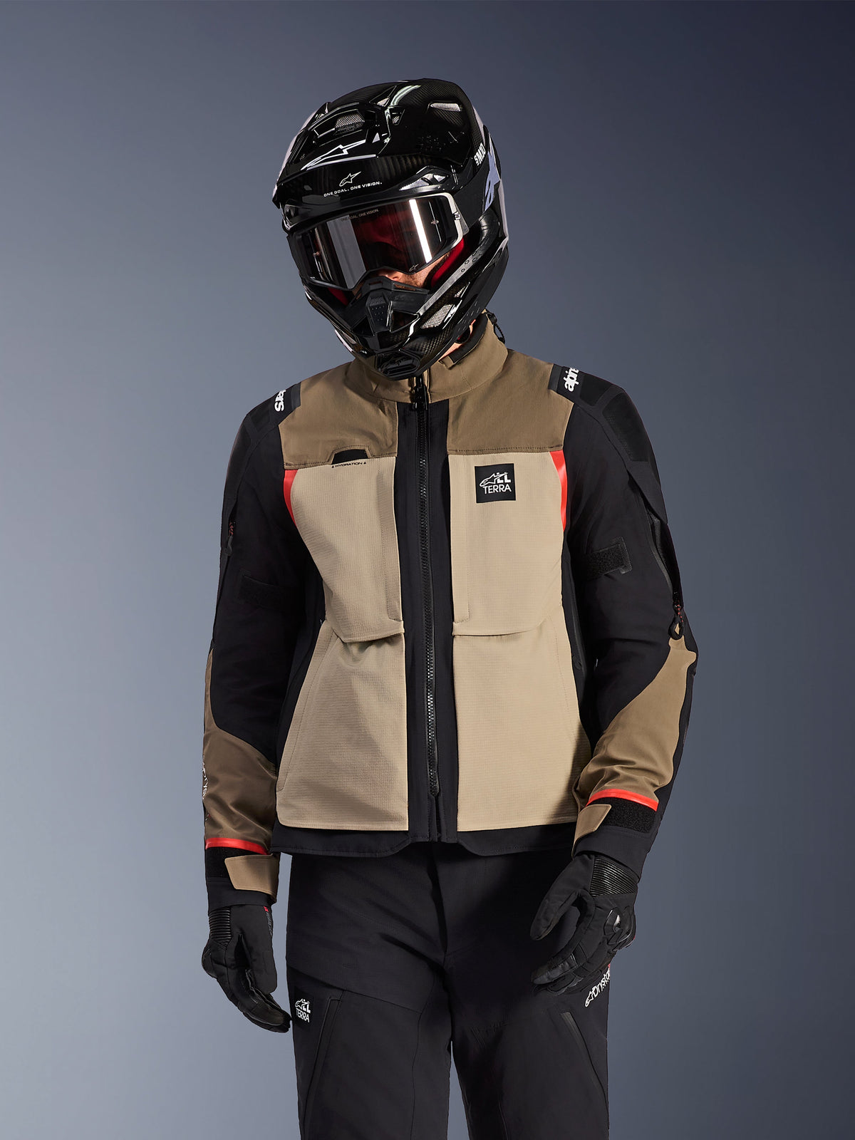 Alpinestars Andes Pro Drystar® XF Jacke, Adventure-Tourenmotorradjacke, Walnut Aluminum Beige und Braun mit schwarzen Einsätzen und roten Akzenten, getragen mit einem schwarzen Alpinestars Helm und Schutzbrille, technische Outdoor-Motorradausrüstung