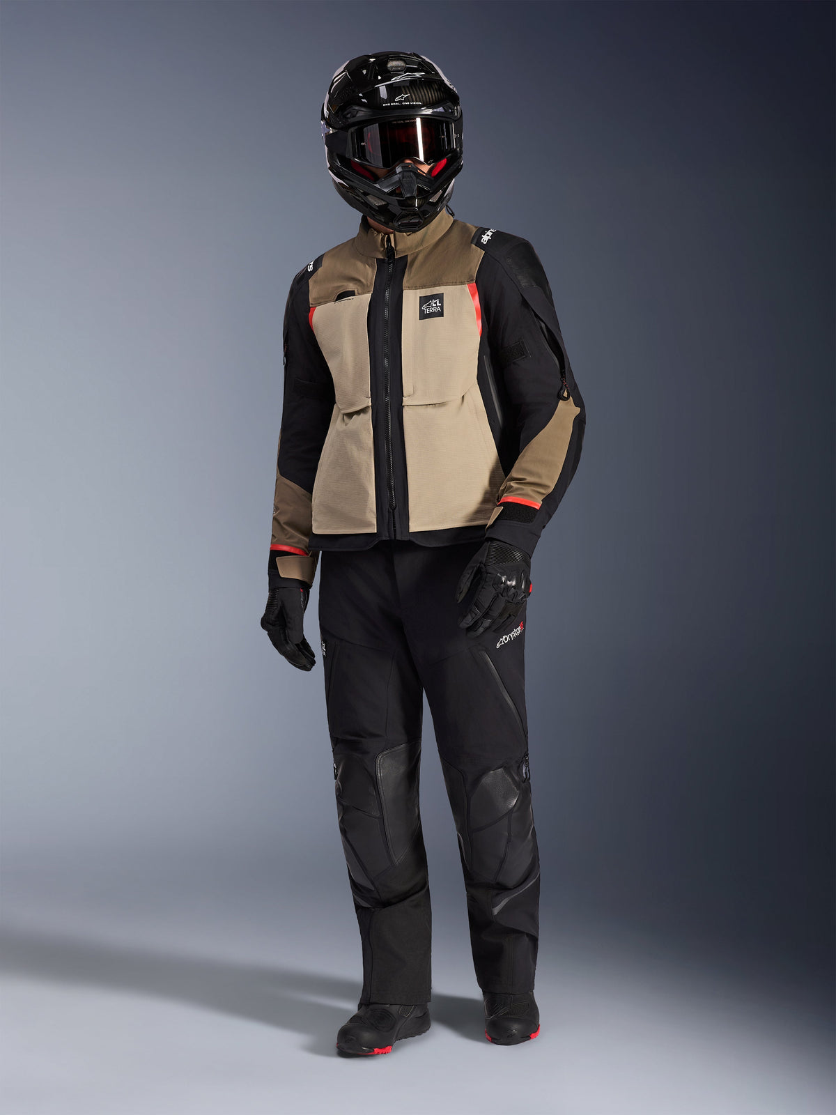 Alpinestars Andes Pro Drystar® XF Jacke in Walnut Aluminum, Adventure-Motorradjacke, getragen mit passender schwarzer Hose, schwarzem Adventure-Helm mit Brille und schwarzen Stiefeln, mit hellbraunen und schwarzen Panels sowie roten Akzenten.