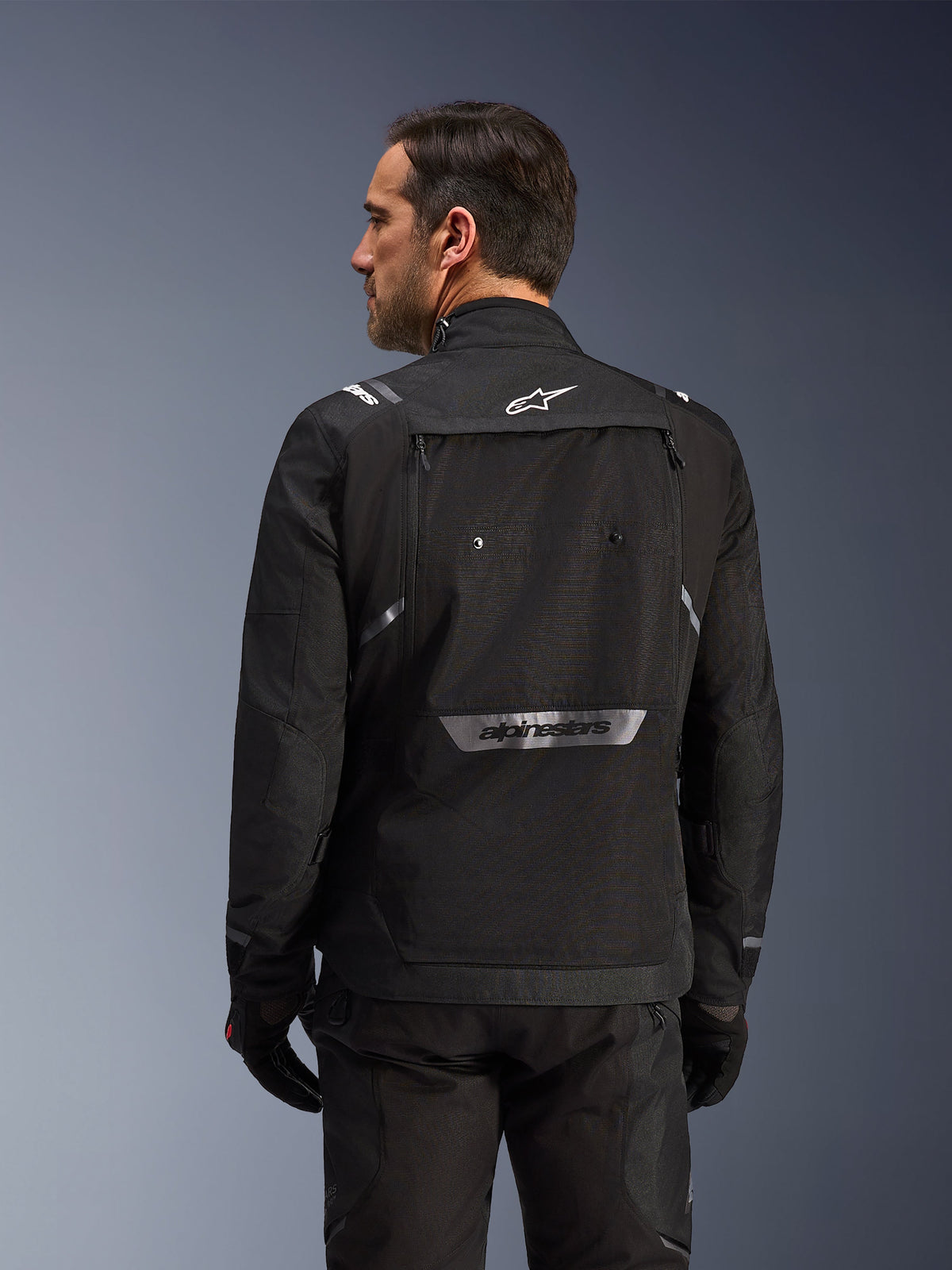 Rückansicht eines Fahrers in der Alpinestars Cusco Drystar® Jacke in Schwarz, eine Textil-Adventure-Tourenjacke mit großer Rückentasche und reflektierendem Branding, kombiniert mit passender schwarzer Hose und Handschuhen
