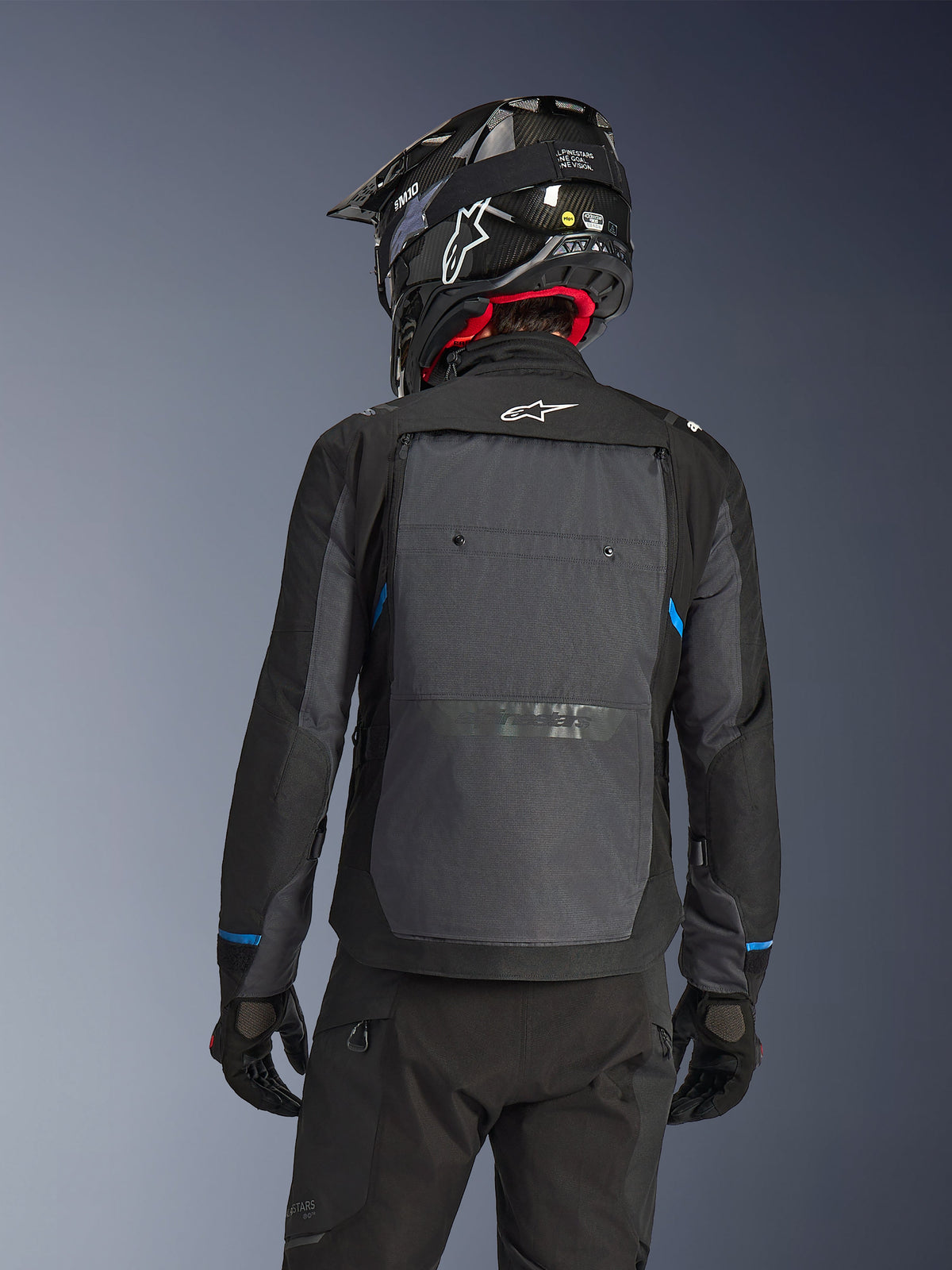 Rückansicht eines Fahrers in der Alpinestars Cusco Drystar® Jacke in Schwarz und Deep Gray, mit einem großen grauen Belüftungspaneel am Rücken und weißem Logo, kombiniert mit einem schwarzen Offroad-Helm und schwarzer Fahrausrüstung für Adventure Touring.