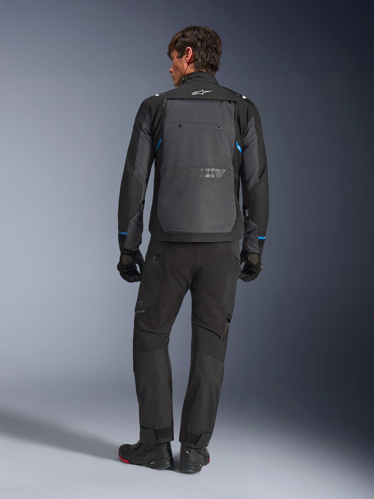 Alpinestars Cusco Drystar® Jacke, Schwarz und Dunkelgrau mit blauen Akzenten, Rückansicht, getragen mit passender schwarzer Adventure-Hose und Handschuhen, mit großem grauen Rückeneinsatz und weißem Logo am Kragen, konzipiert für Straßen- und Adventure-Touren
