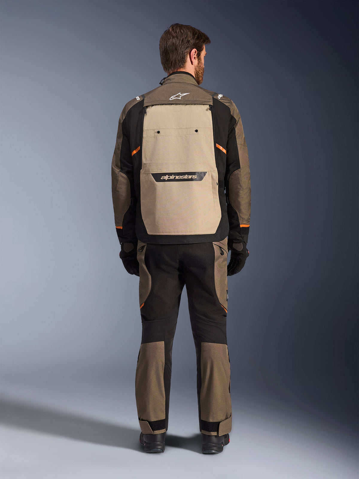 Alpinestars Cusco Drystar® Jacke, Rückansicht, Walnut Aluminum Black, Textil-Adventure-Touring-Ausrüstung, großes beiges Belüftungspanel am Rücken mit schwarzem Branding, orangefarbene Akzente, getragen mit passender Hose und Handschuhen