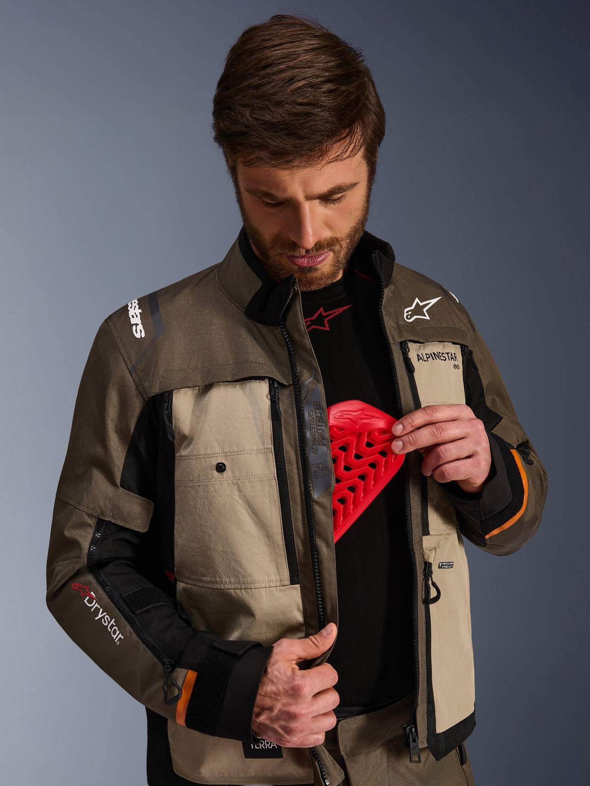 Alpinestars Cusco Drystar® Jacke, Walnut Aluminum Black, getragen von einem Mann, der die Brustprotektorentasche mit einem roten Protektoreinsatz zeigt; Textil-Motorradjacke mit Belüftungsöffnungen mit Reißverschluss und Branding für Adventure-Touring.