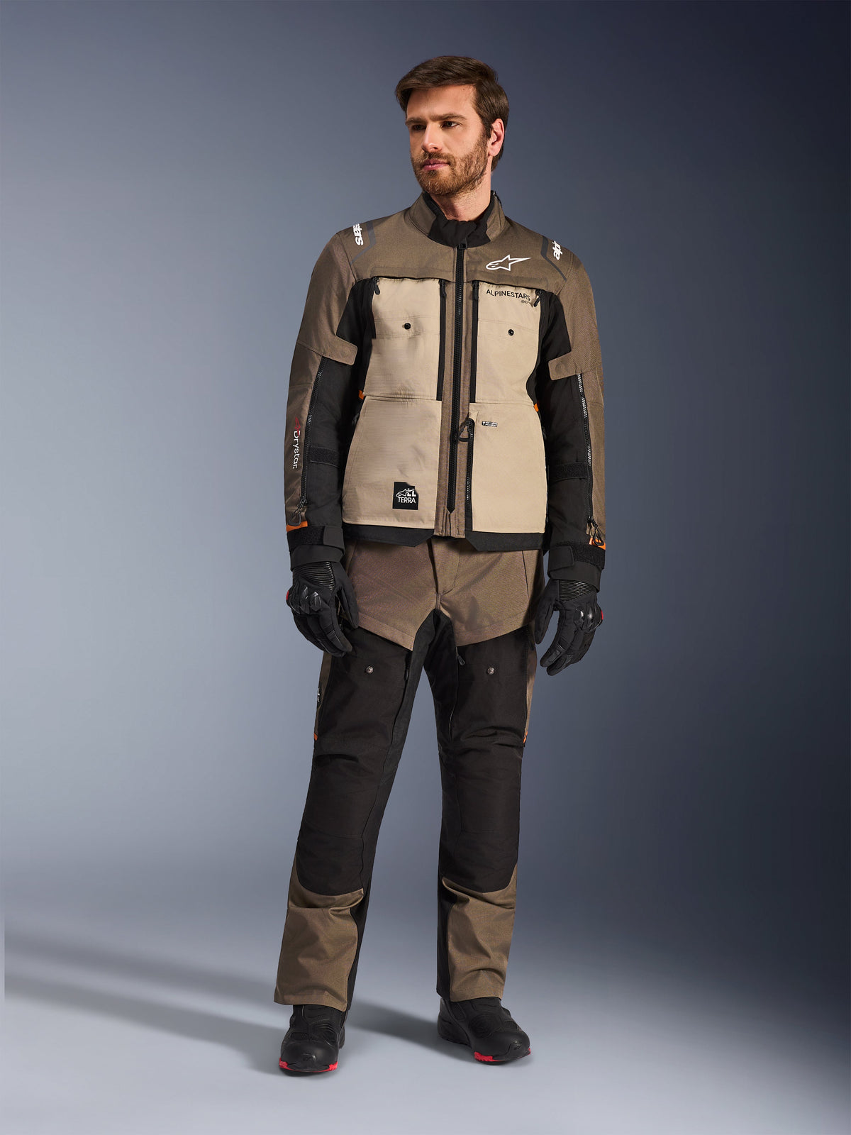 Alpinestars Cusco Drystar® Jacke in Walnut Aluminum Black, getragen von einem männlichen Fahrer mit passender Adventure-Tourenhose, schwarzen Handschuhen und Stiefeln, mit beigen Brustpaneelen, schwarzen Ärmeln mit dezenten orangefarbenen Akzenten und funktionellen Belüftungsreißverschlüssen für Motorrad-Straßenfahrten.