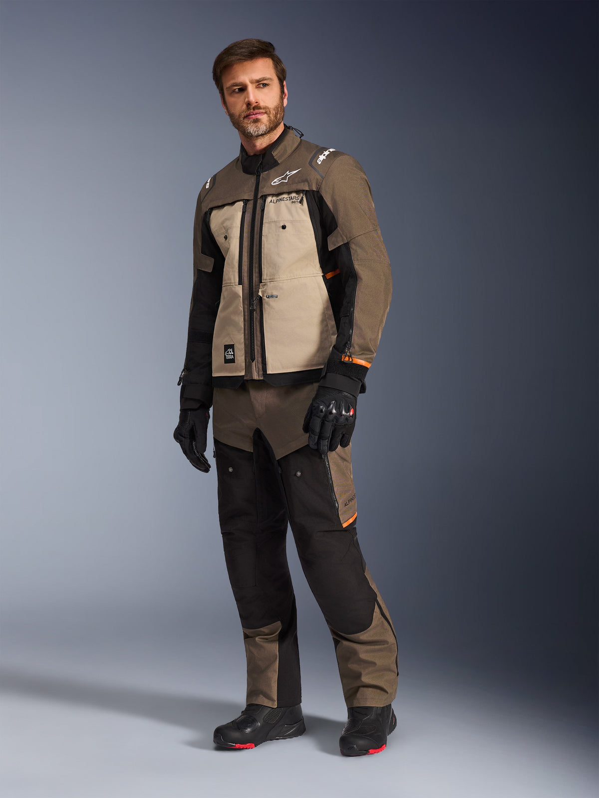 Alpinestars Cusco Drystar® Jacke in Walnut Aluminum Black, getragen von einem männlichen Model mit passender Motorradhose und Handschuhen, Adventure-Touring-Design mit hellbraunen Brustpaneelen und schwarzen Akzenten.