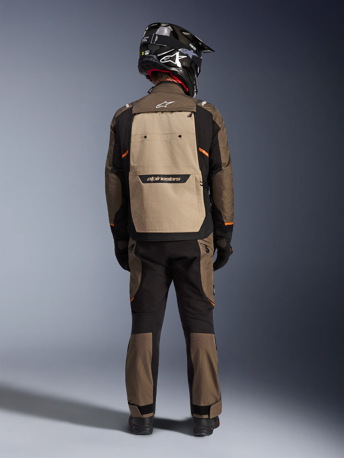 Fahrer in der Rückansicht mit der Alpinestars Cusco Drystar® Jacke in Walnut Aluminum Black, große aluminiumfarbene Rückentasche und orangefarbene Akzente, passende Adventure-Hose, schwarzer Offroad-Helm und Handschuhe, strapazierfähige Textil-Motorradausrüstung für Touren