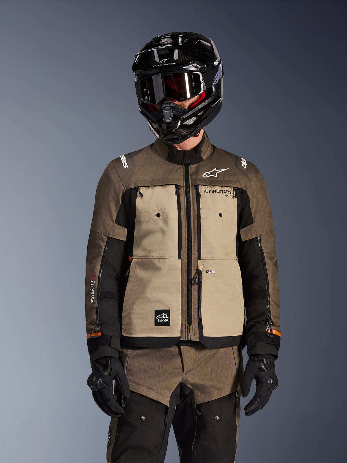 Fahrer in der Alpinestars Cusco Drystar Jacke in Walnut Aluminum Black, Adventure-Touring-Motorradausrüstung mit schwarzem Helm, Brille und schwarzen Handschuhen, Frontansicht der technischen Textilkonstruktion mit Taschen und Branding.
