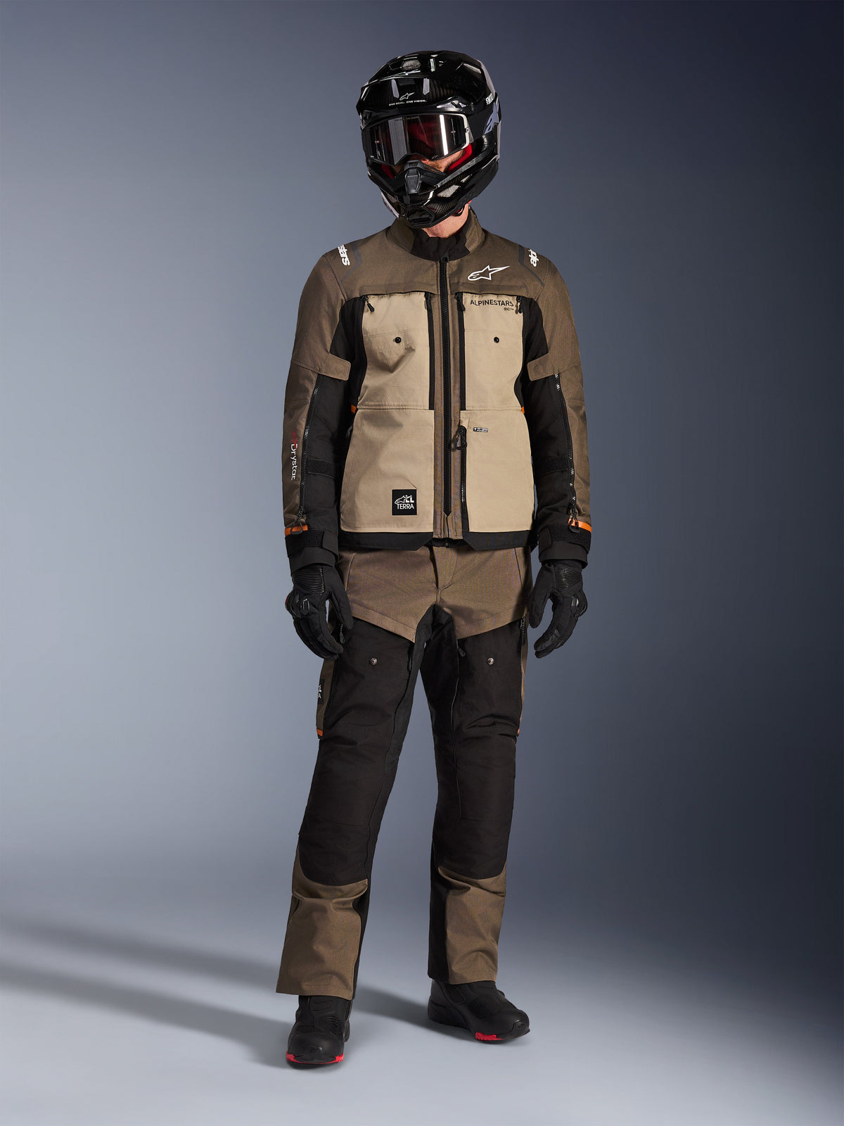 Alpinestars Cusco Drystar® Jacke, Walnut Aluminum Black, Adventure-Touring-Motorradjacke getragen mit passender Hose und einem schwarzen Helm, mit hellbraunen Brustpaneelen und braunen Ripstop-Ärmeln, technische Allwetter-Fahrausrüstung