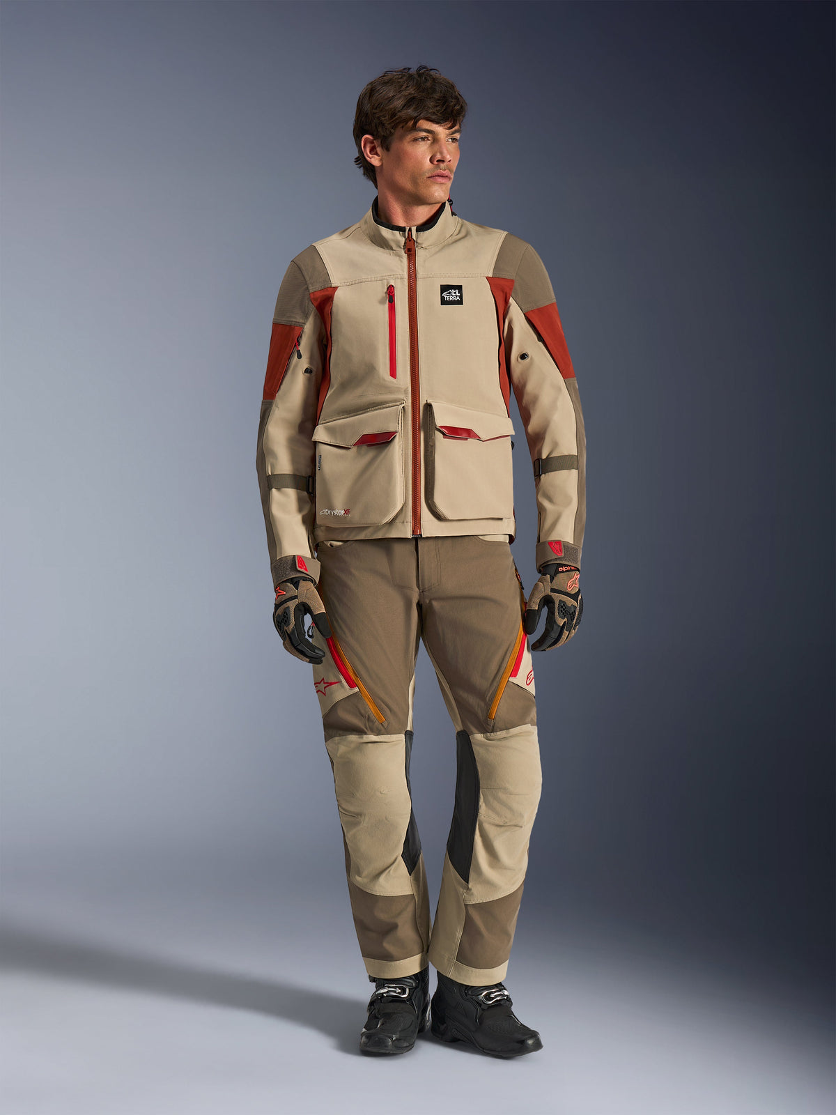 Maxdura Drystar®XF Laminated Armored Shell, Adventure-Motorradjacke in Aluminum Walnut Rust Brown, getragen von einem männlichen Model mit passender Adventure-Hose, Handschuhen und Stiefeln, Ganzkörper-Studioaufnahme einer professionellen Tourenausrüstung.