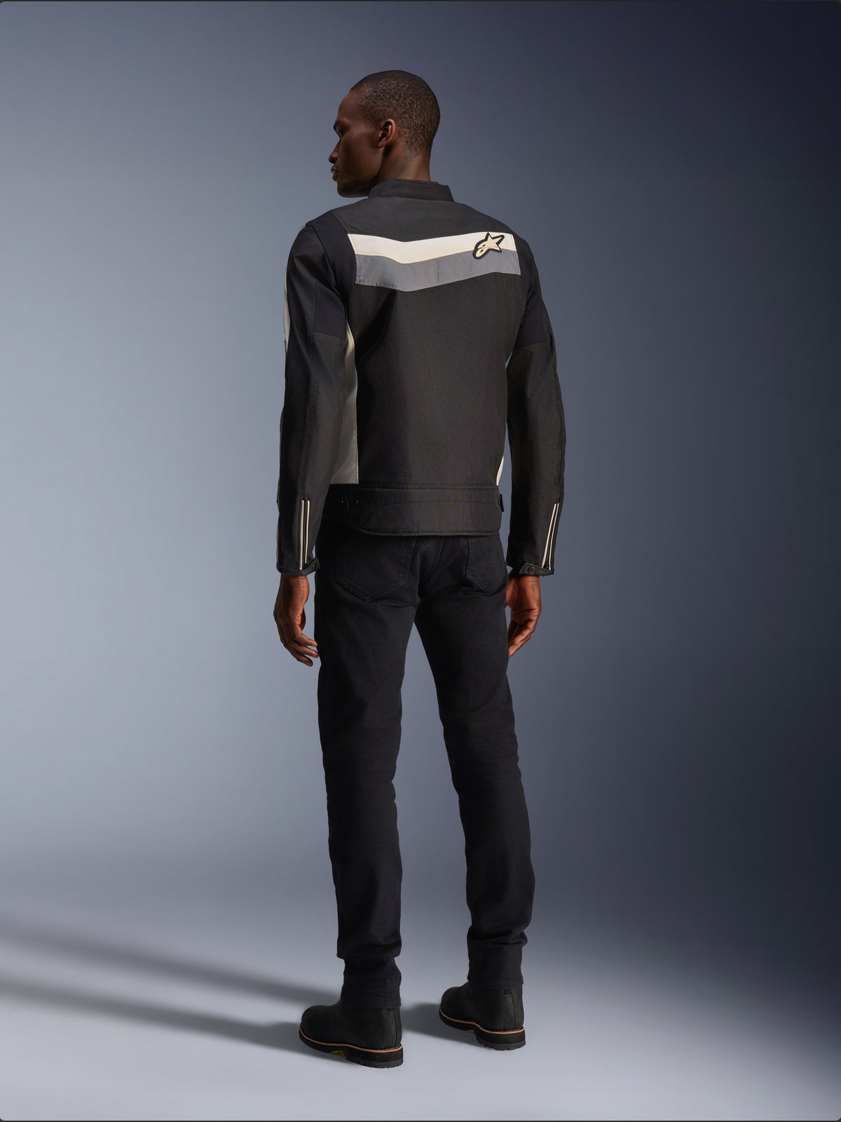 Männliches Model von hinten in der Alpinestars T-Dyno WR Jacke in Black Dark Gray Ecru, Textil-Motorradjacke mit horizontalem Streifen und Stern-Logo auf dem Rücken, kombiniert mit schwarzer Motorradhose und Stiefeln, urbane Motorradausrüstungs-Ästhetik.