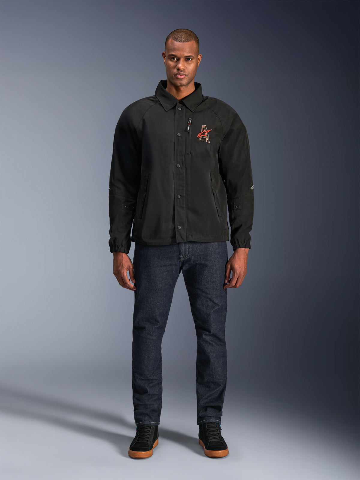 Alpinestars Revved WR Jacke in Schwarz, getragen von einem Modell mit dunkelblauen Jeans und schwarzen Sneakern, Vorderansicht, mit Druckknopfverschluss und einem 3D-Stickerei-Logo auf der Brust, moderne Urban-Motorradausrüstung.