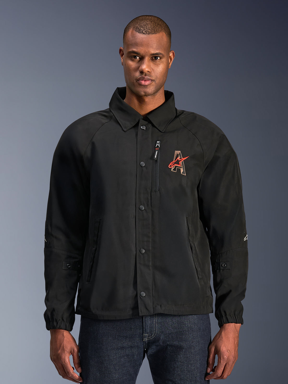 Alpinestars Revved WR Jacke, schwarze Motorradjacke mit Hemdkragen und Druckknopfleiste, rote 'A' Logo-Stickerei auf der Brust, vertikale Brusttasche mit Reißverschluss, elastische Bündchen, urbanes Coach-Jacken-Design