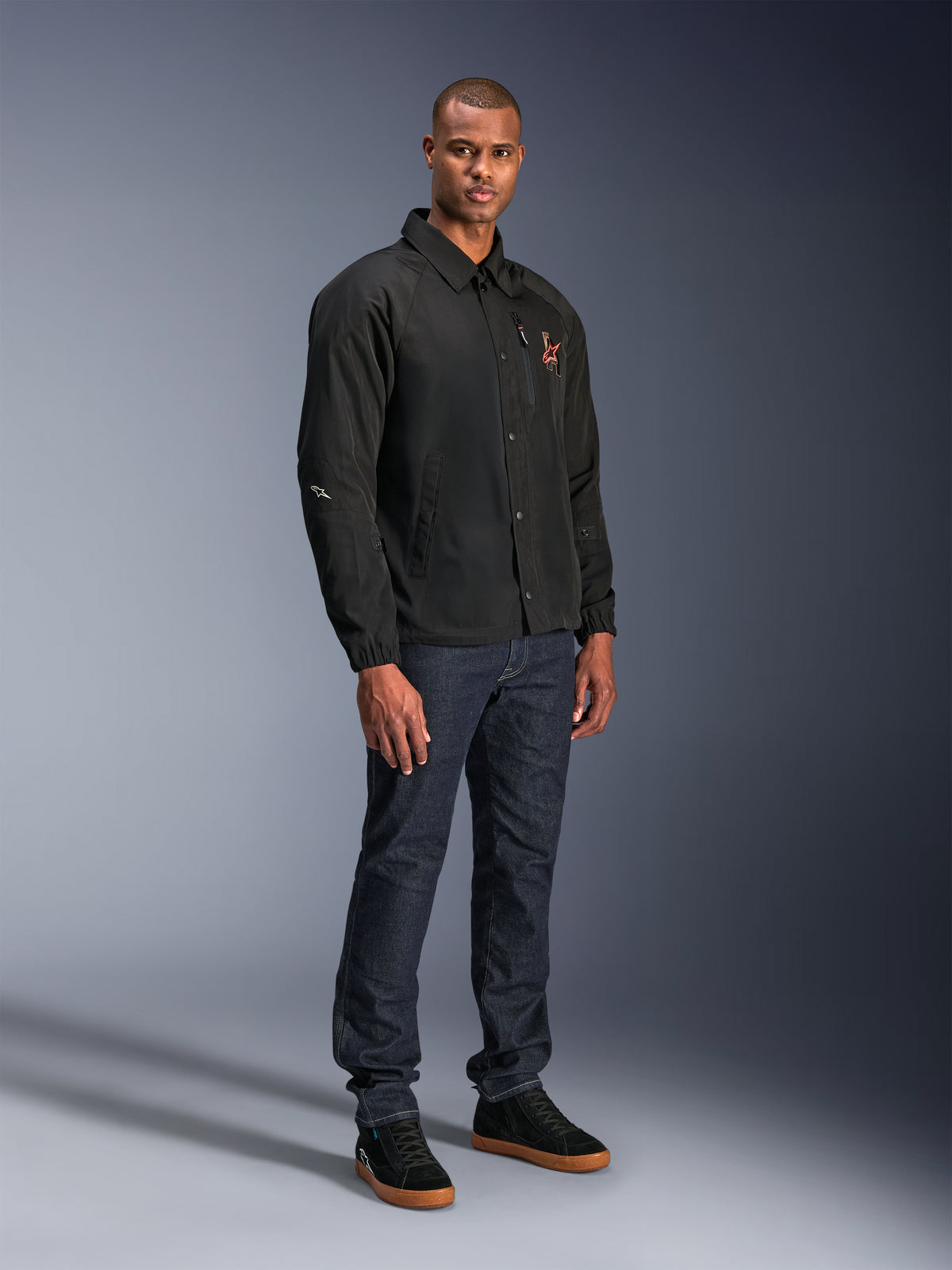 Alpinestars Revved WR Jacke in Schwarz, Motorradjacke im urbanen Coach-Stil, getragen von einem Model mit dunkelblauen Jeans und schwarzen Sneakern, mit Druckknopfleiste vorne und gesticktem Logo, minimalistisches und sportliches Design für Straßenfahrten