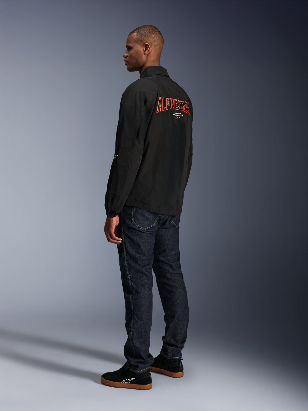 Alpinestars Revved WR Jacke, Schwarz, mit großem orangefarbenem 3D-gesticktem Alpinestars-Branding auf dem Rücken, Dreiviertel-Rückansicht eines Models in dunkelblauen Jeans und schwarzen Sneakern, moderne urbane Motorradjacke mit Coach-Jacken-Silhouette