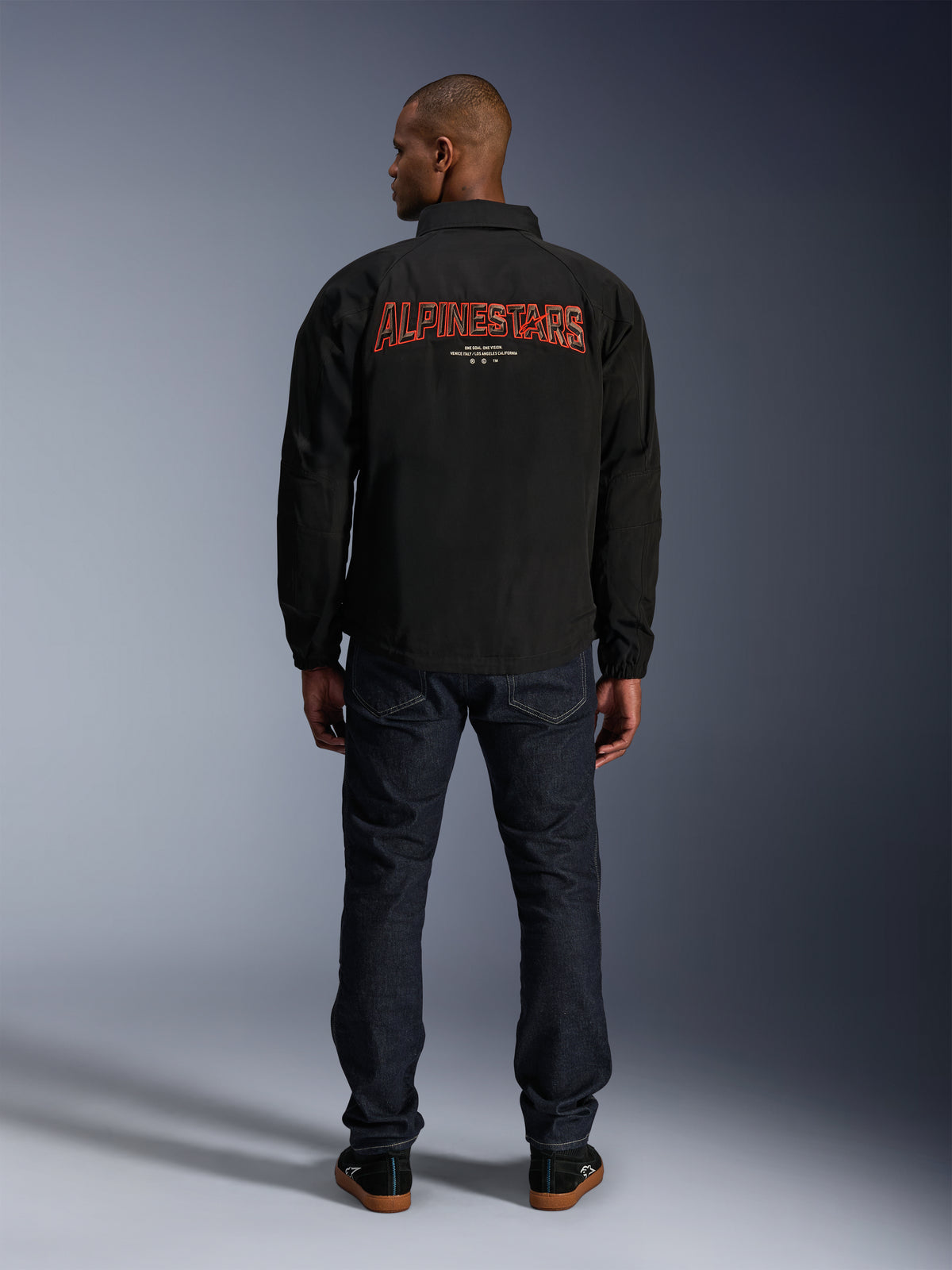 Alpinestars Revved WR Jacke, Schwarz, Rückansicht mit großer roter 'ALPINESTARS' Stickerei im 3D-Effekt, urbane Jacke im Coach-Stil, getragen mit dunkler Denim-Jeans und schwarzen Sneakern, minimalistische Motorradbekleidung